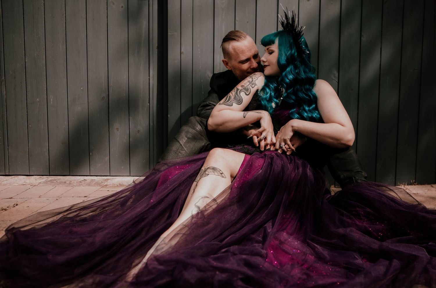Couple in gothic wedding attire, woman in purple gown with teal hair, man with tattoos, sitting together against wooden wall.