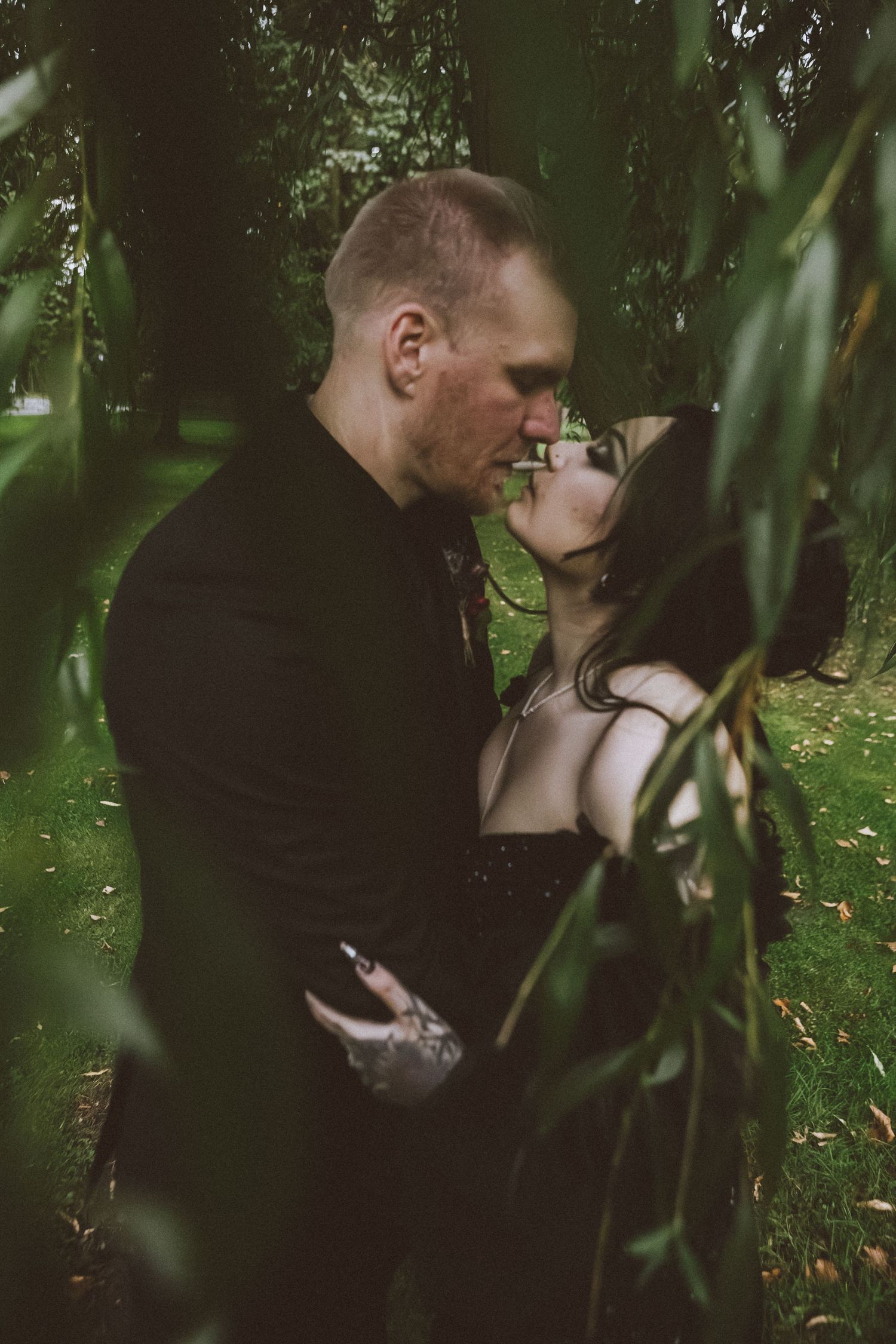 A couple shares a romantic kiss among weeping willow branches in a lush green park setting.