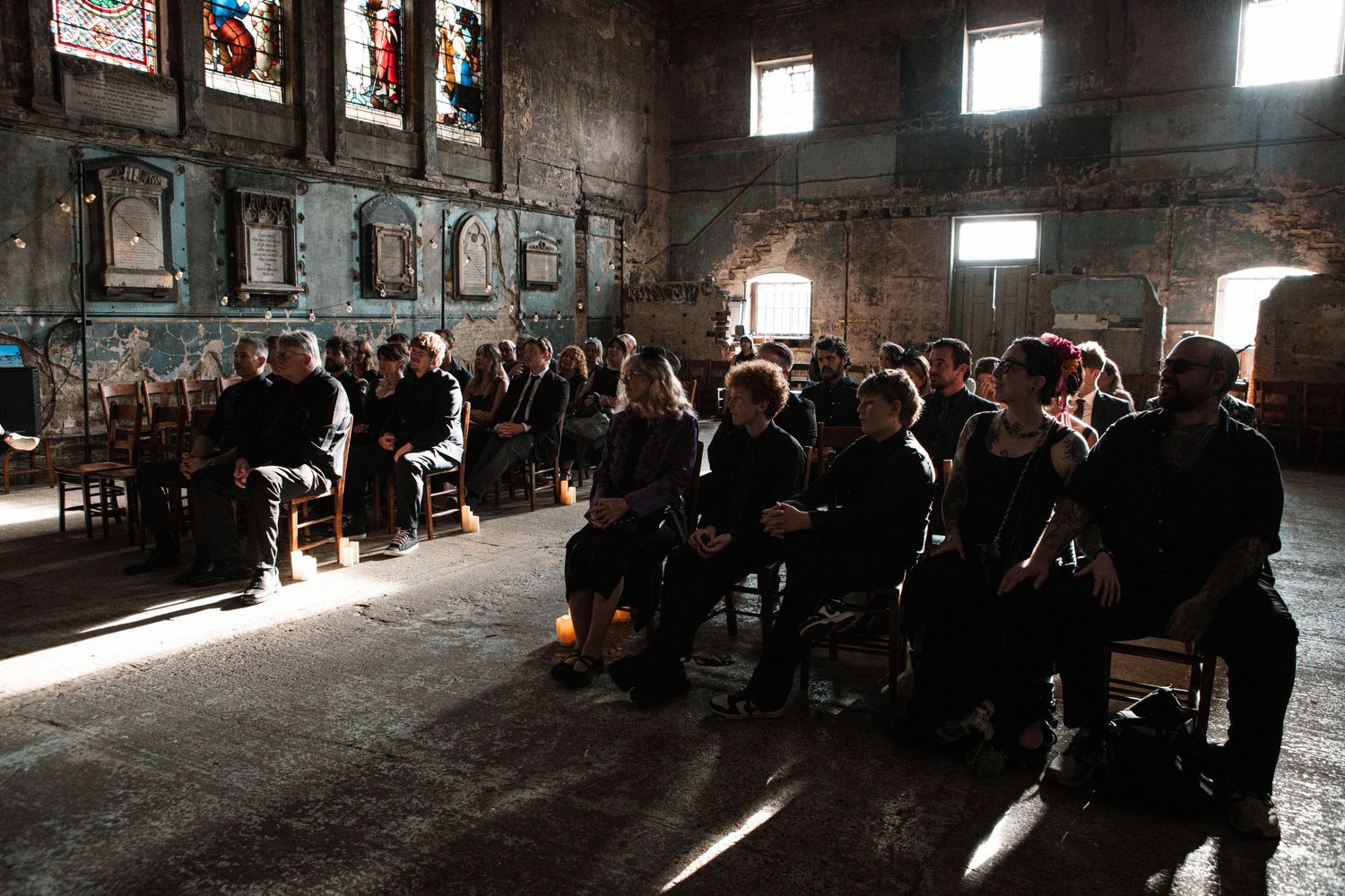 Audience seated in rows inside a rustic stone church with stained glass windows during an indoor event.