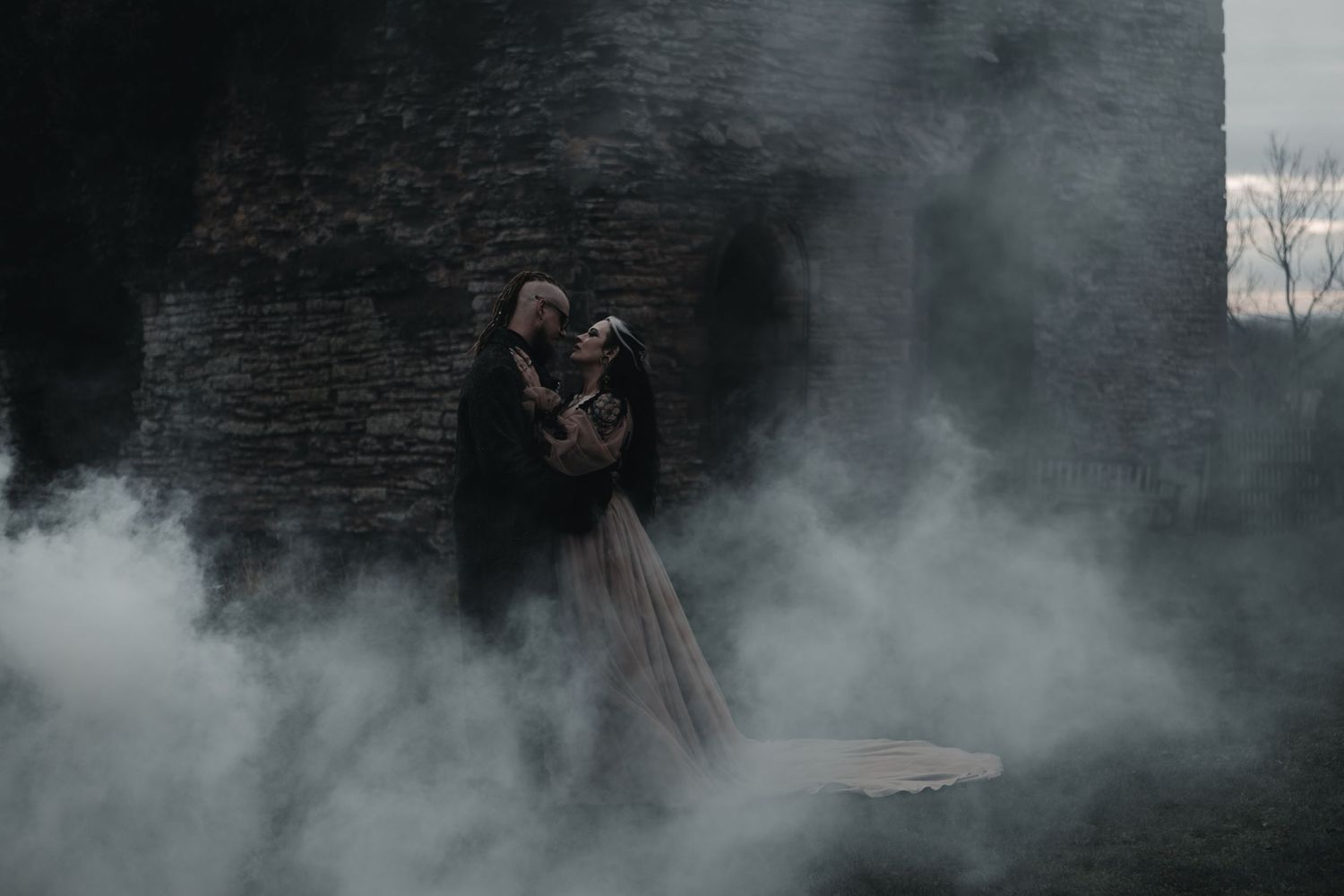 Couple embracing in misty fog near stone ruins for a dark gothic wedding photoshoot.