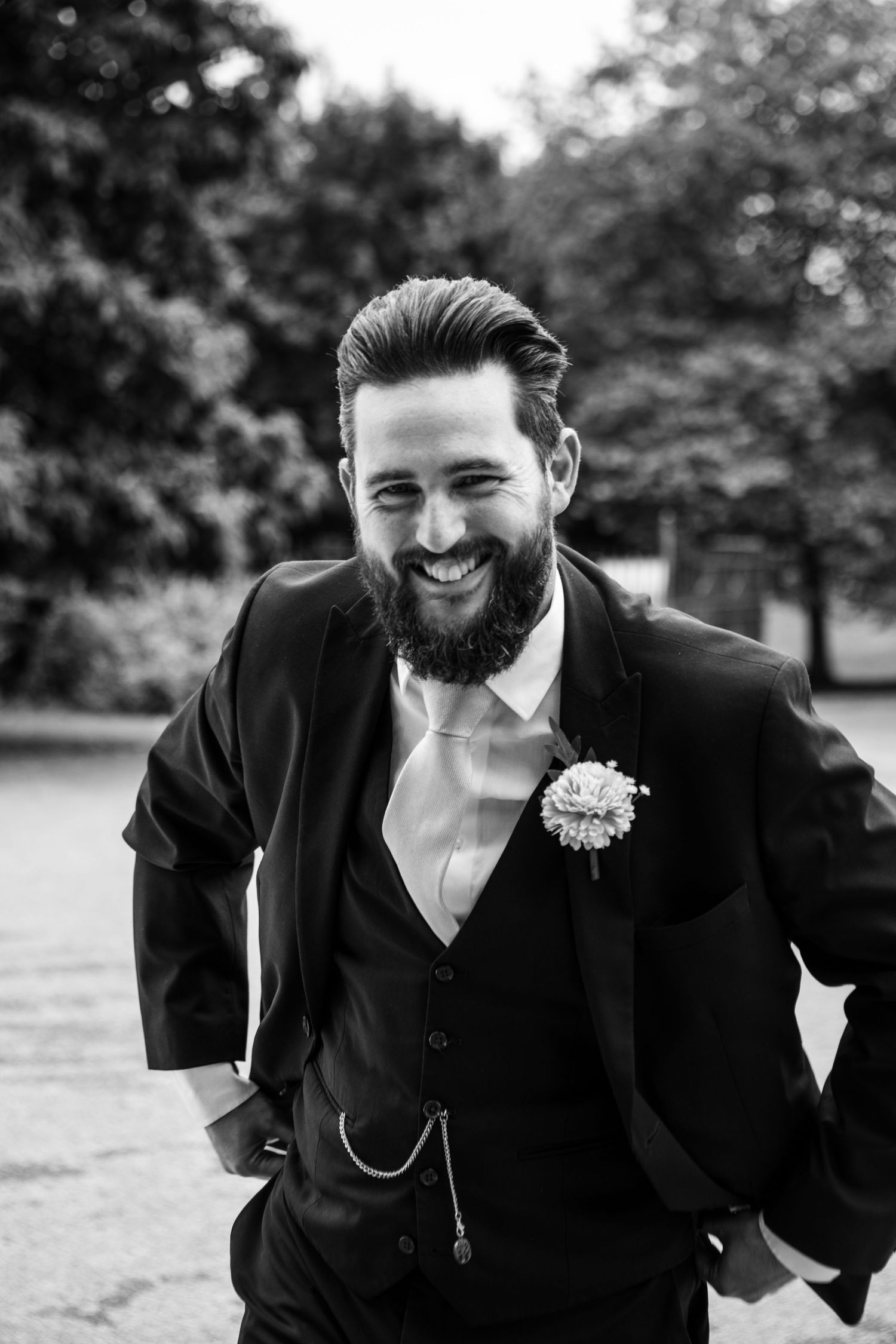 Groom in classic black three-piece suit and boutonniere shows candid expressions in outdoor photo series.