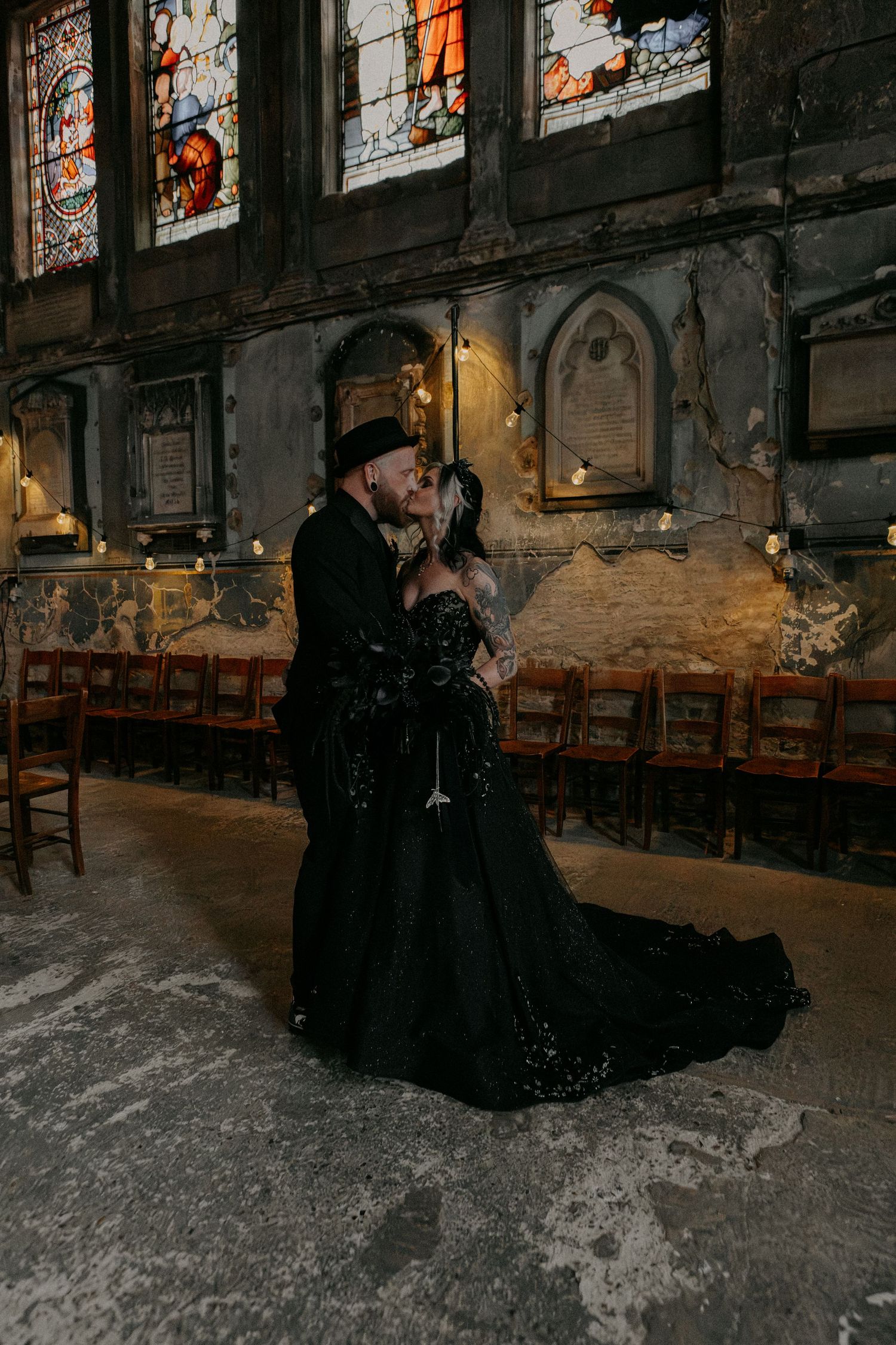 Couple in matching black wedding attire share a romantic embrace inside a gothic church with stained glass windows.