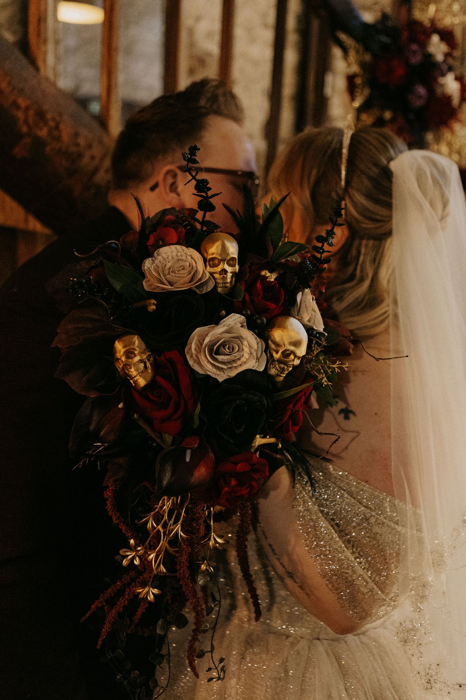 Gothic wedding bouquet with gold skulls, dark red and white roses, black flowers, held by bride in glittery gown with veil.