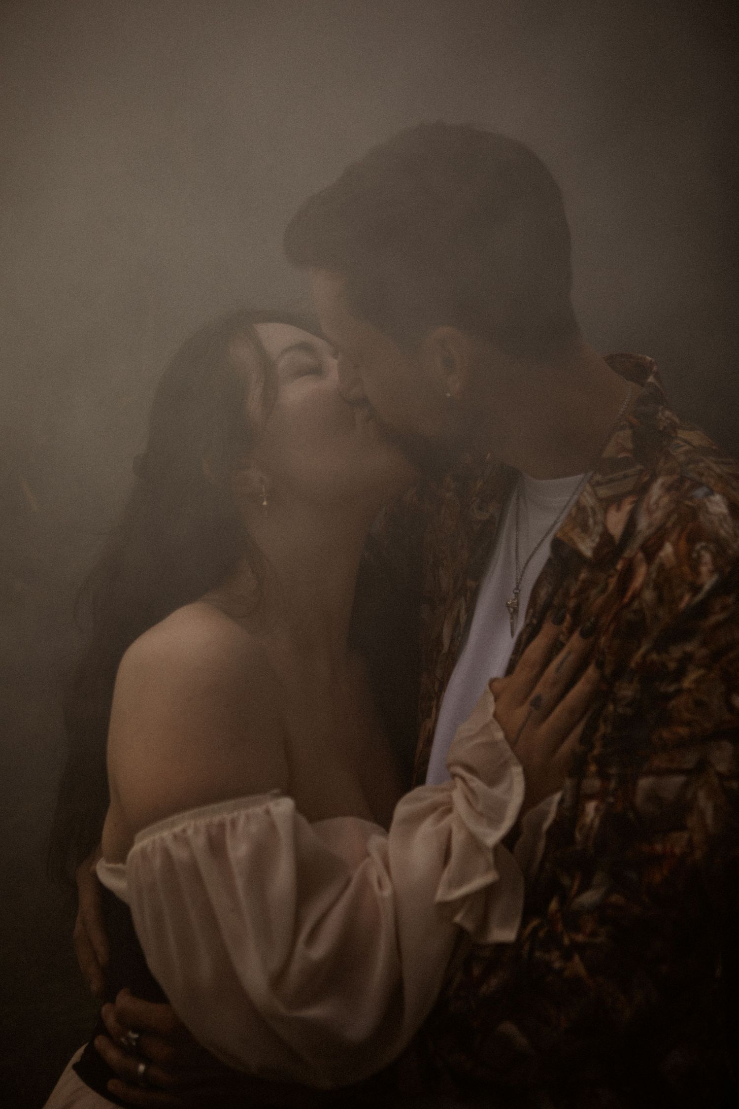 A romantic couple embraces in a misty setting, with the woman wearing an off-shoulder white dress and the man in dark attire.