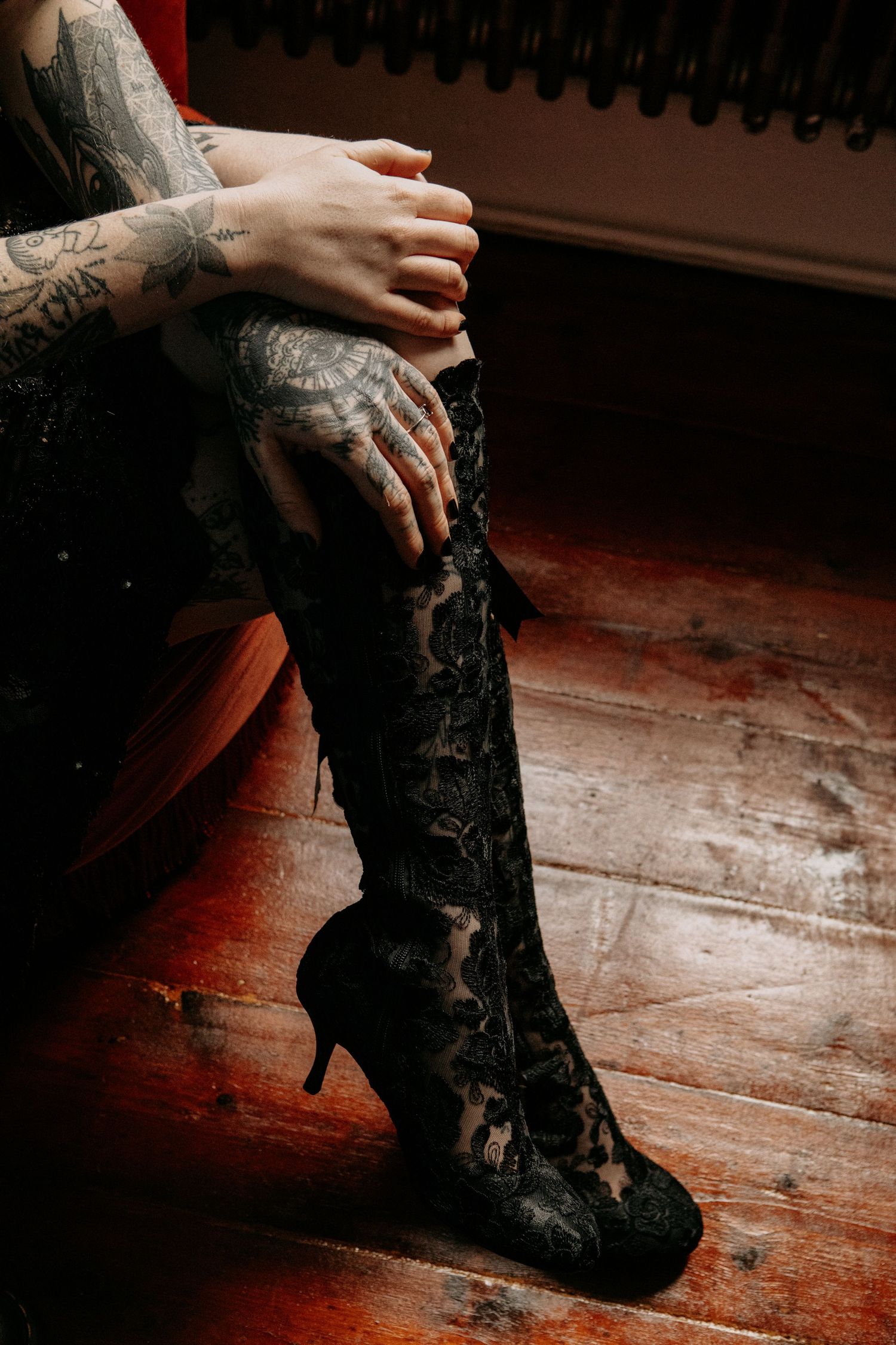 Tattooed hands pulling on a black lace knee-high heeled boot over tattooed legs.