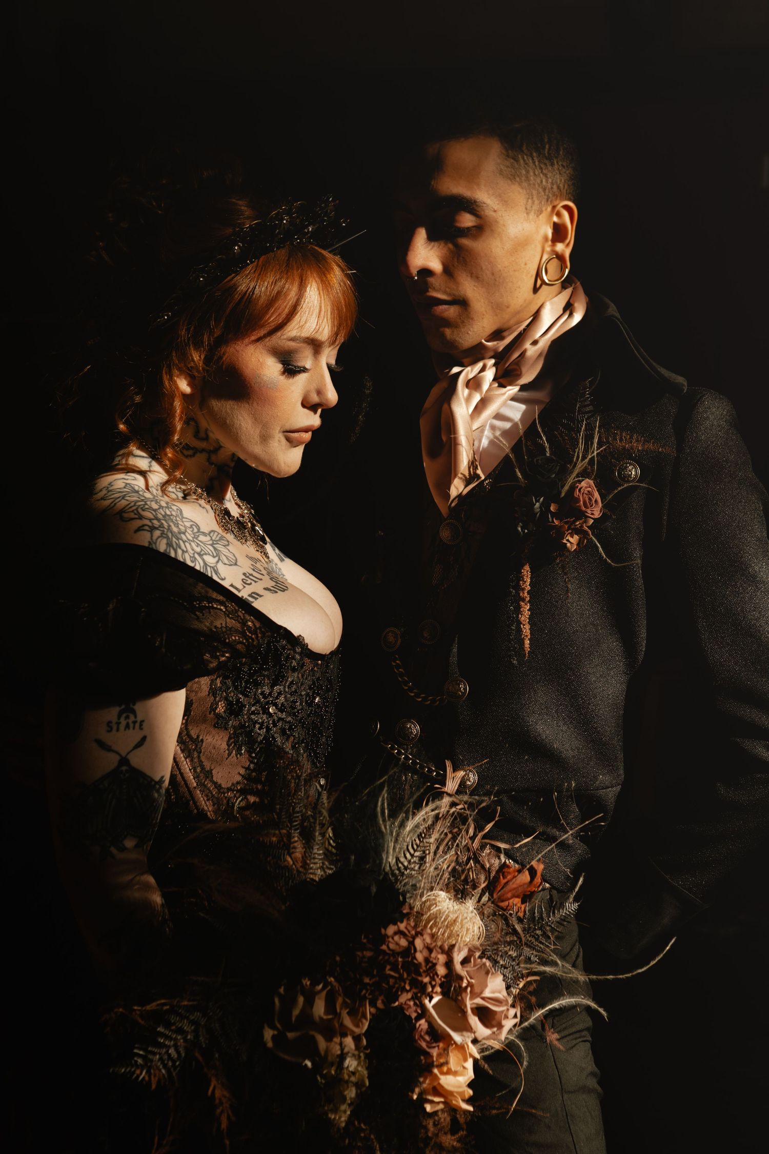 Gothic bride and groom in elegant dark wedding attire posing against a black backdrop.