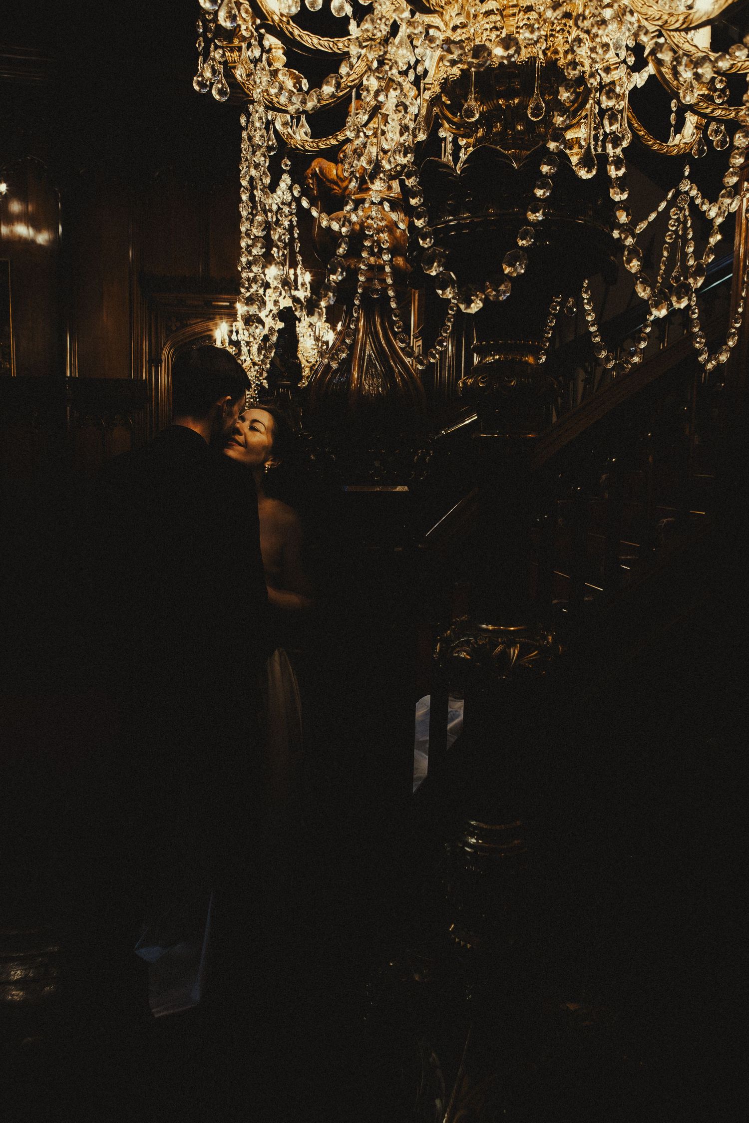 Couple embracing romantically under a glittering crystal chandelier in a dimly lit elegant venue.