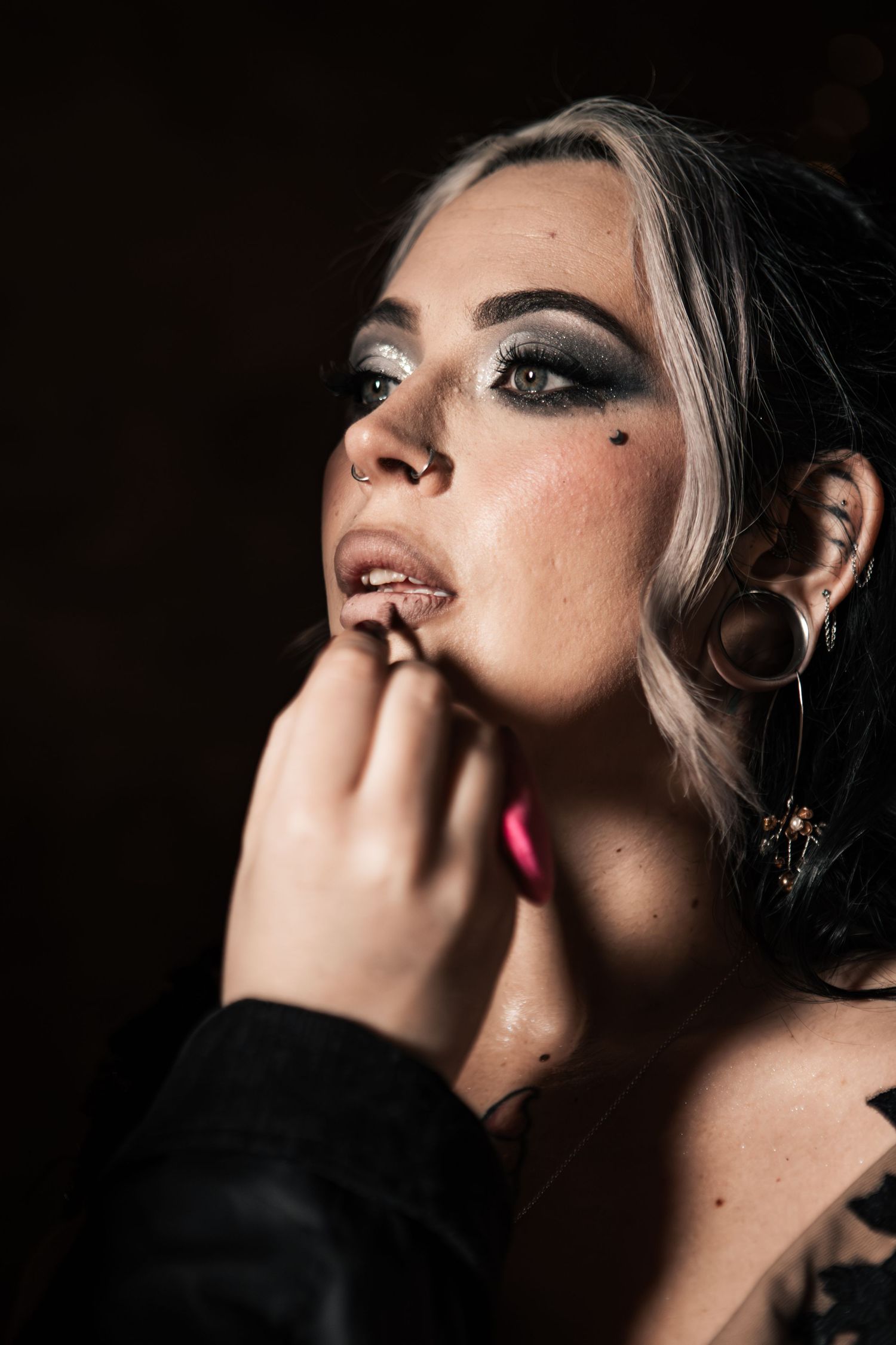 A woman with dramatic smoky eye makeup, gauge earrings, and dark lipstick poses in moody low-key lighting.