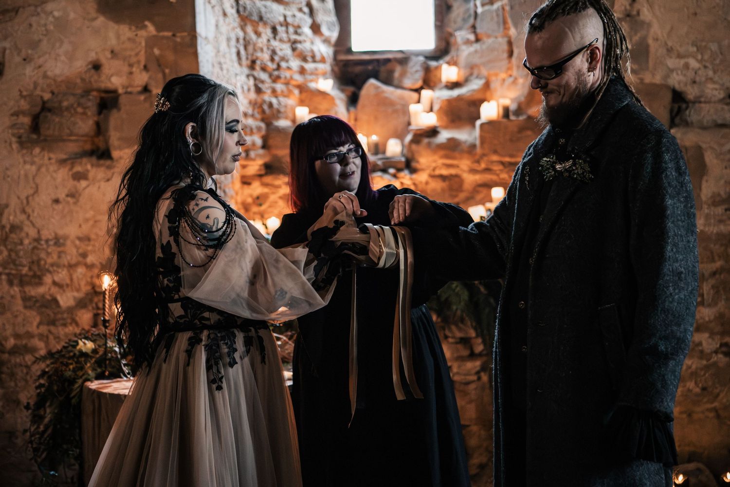 Gothic wedding officiant in black robe performing hand-fasting ritual with bride and groom in candlelit stone chapel.