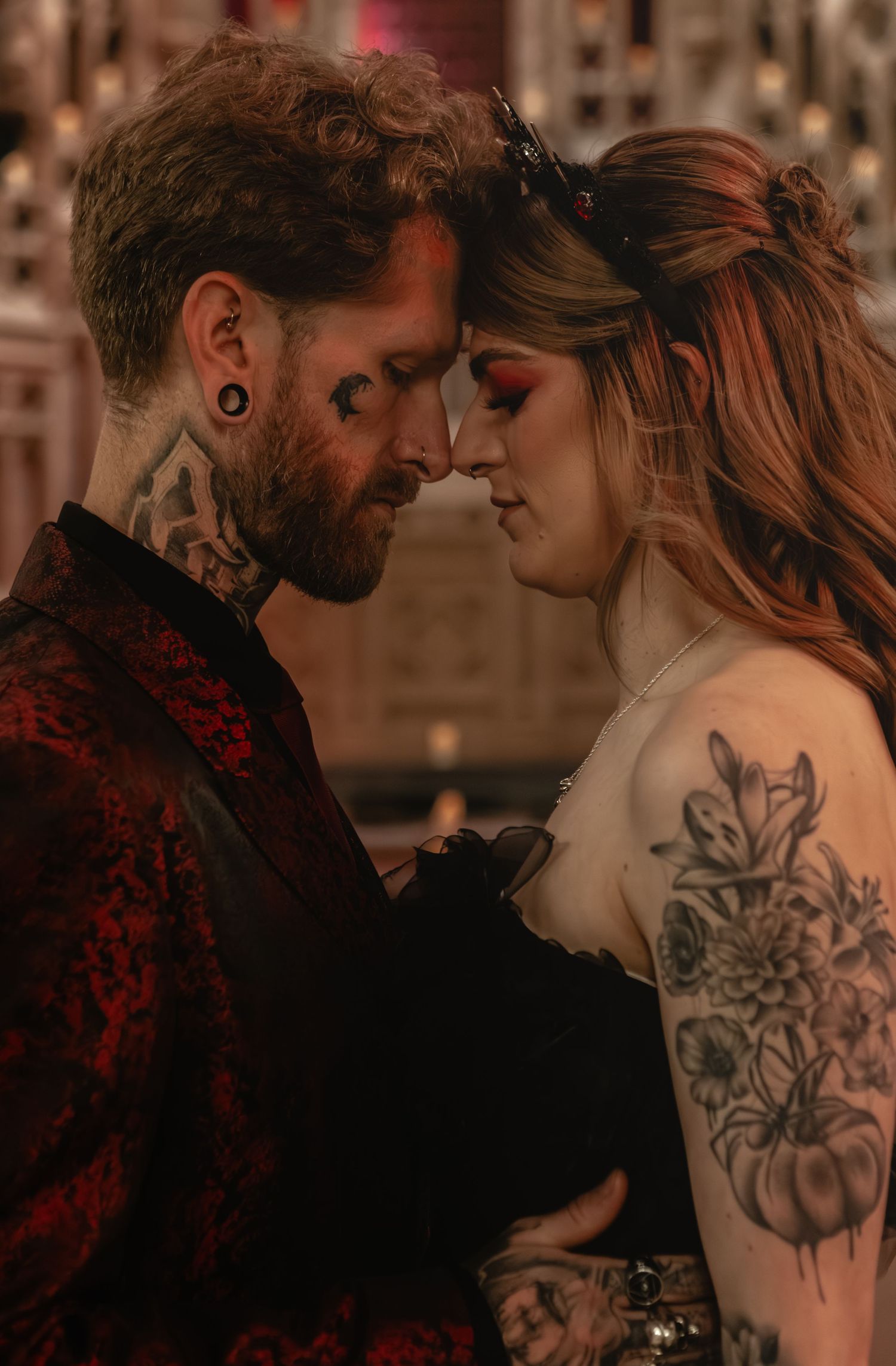 A tattooed couple in elegant attire share an intimate forehead-to-forehead moment in a romantic, dimly lit setting.