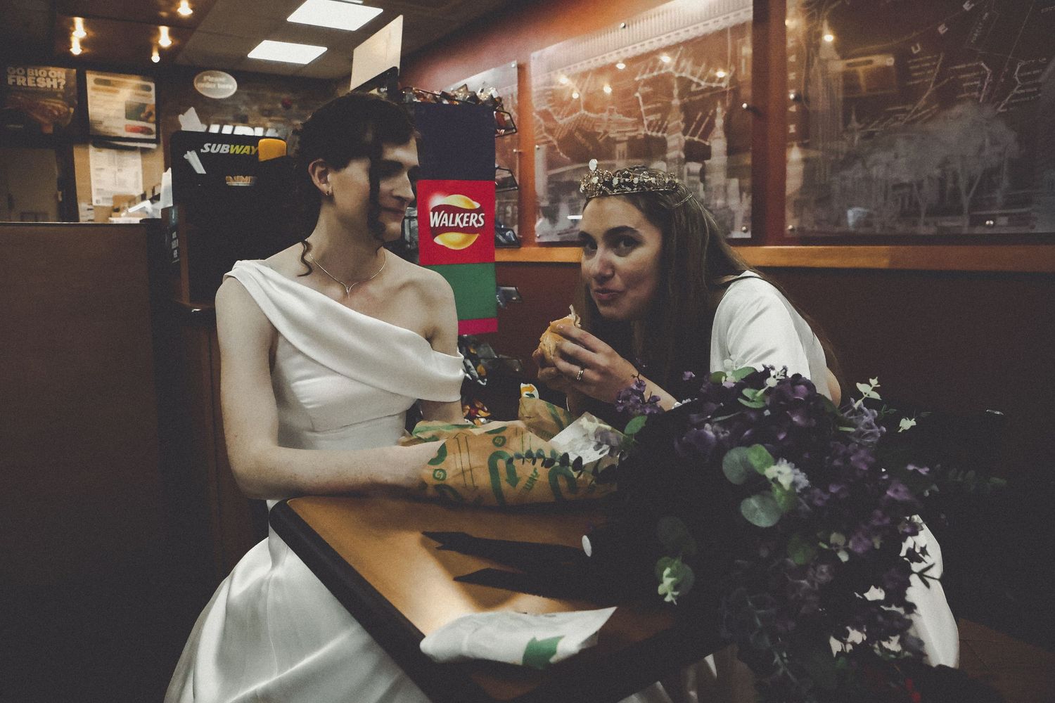 Two brides in white wedding dresses share a meal at a diner booth, one wearing a floral crown holding a purple bouquet.