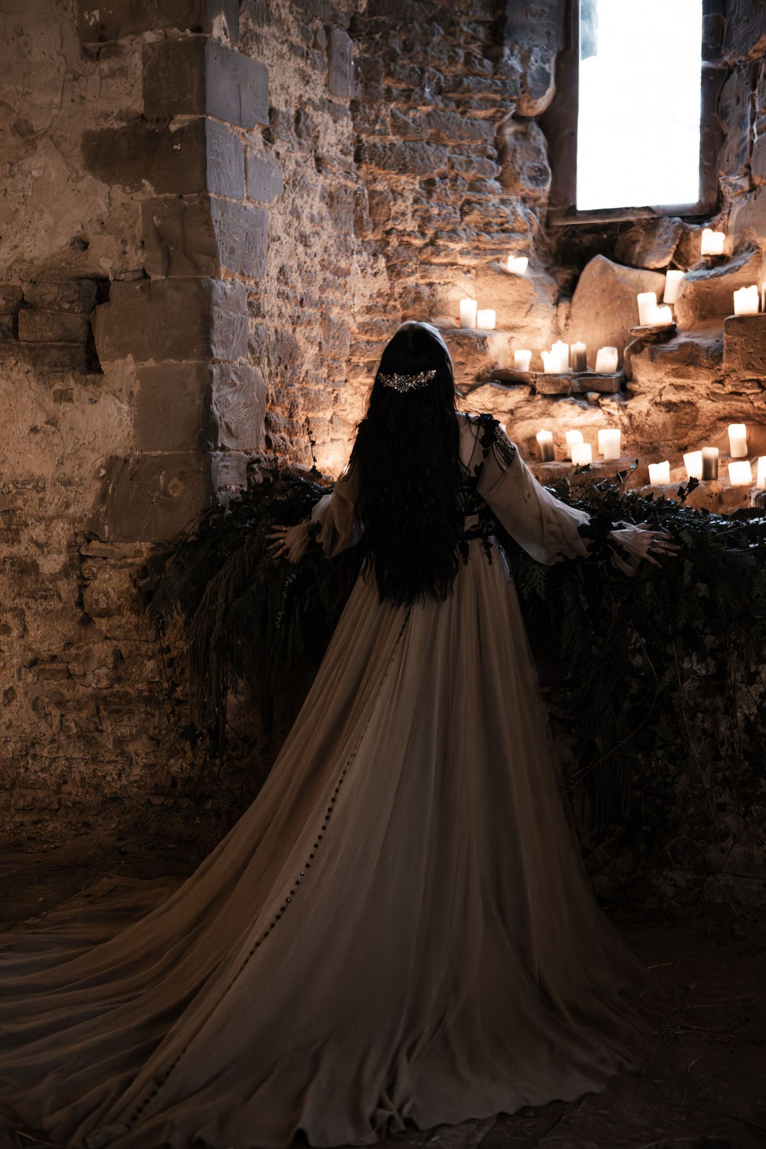 Woman in elegant medieval gown with long dark hair stands before candlelit stone wall in ancient castle.