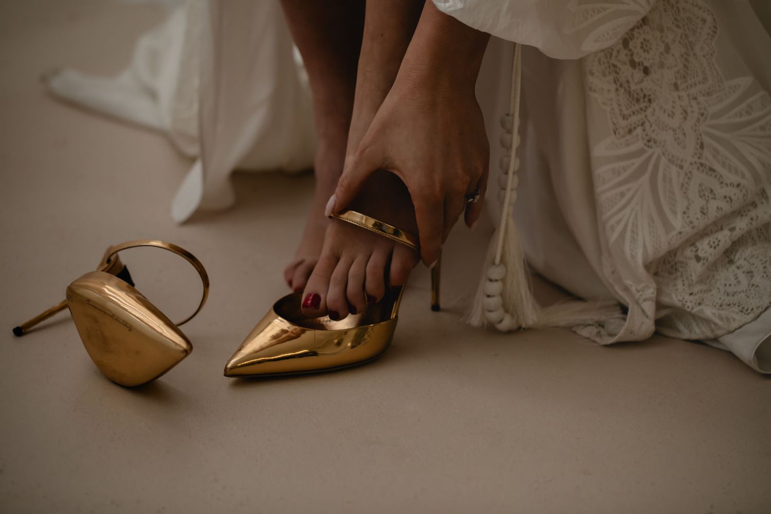 Close-up view of someone putting on a metallic gold bridal shoe next to a matching clutch purse on a beige floor.