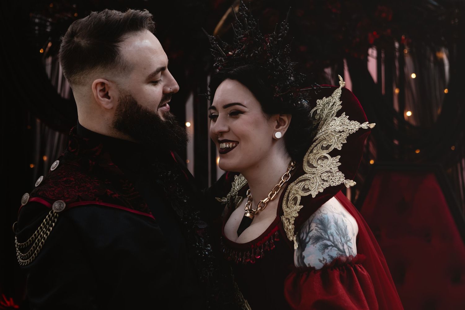Couple in elaborate Renaissance-inspired wedding attire smiling at each other in dramatic red and black costumes.