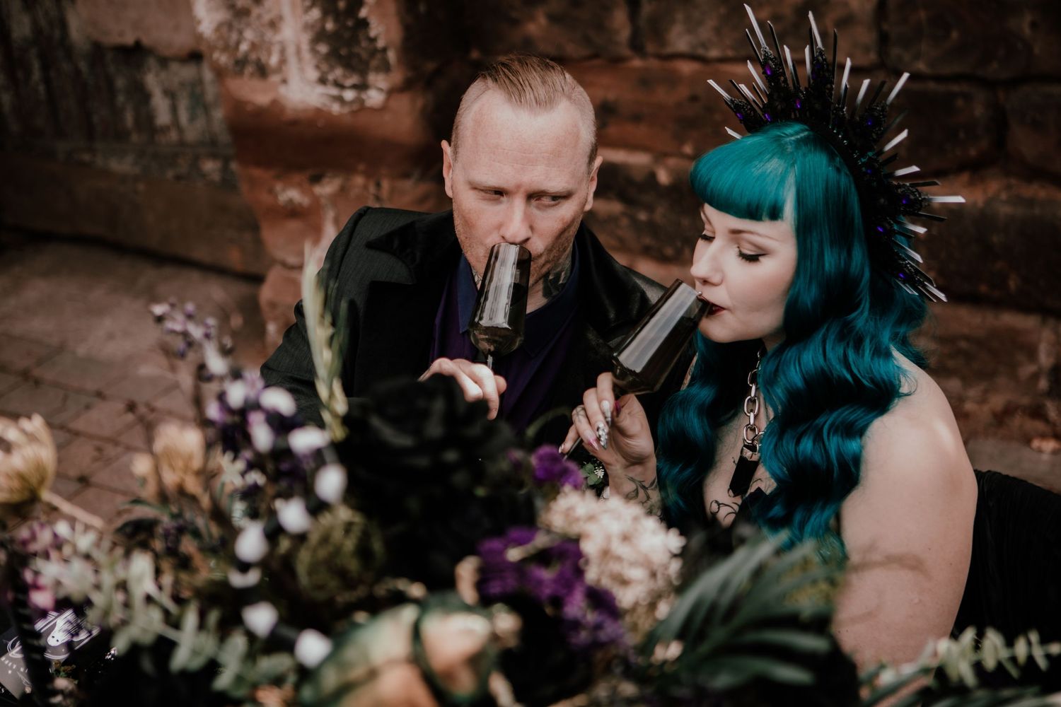 Alternative wedding couple sharing a drink, bride with teal hair and punk mohawk, groom in black suit, floral arrangement.