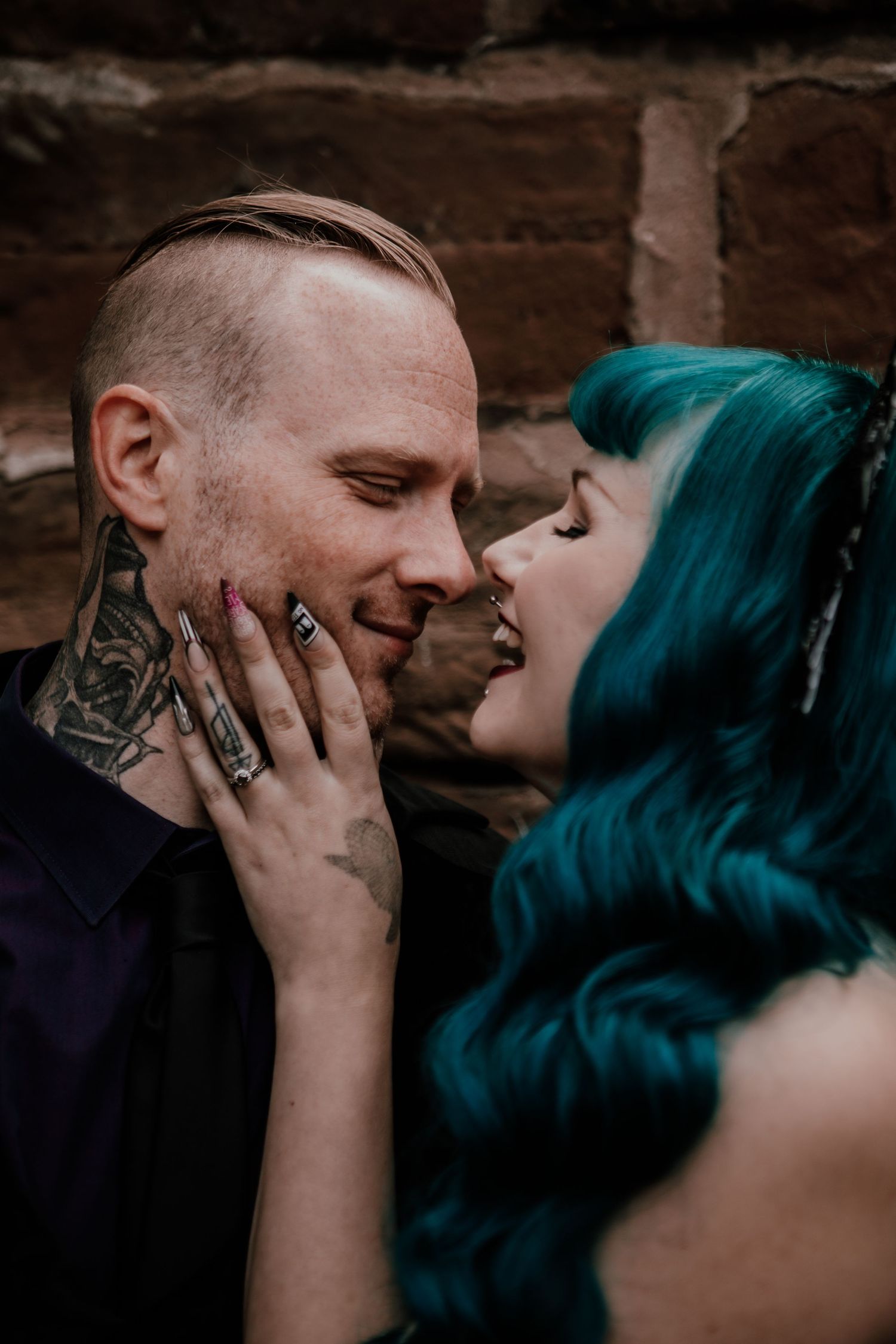 Tattooed couple with teal-haired woman and shaved-head man sharing an intimate moment against a brick wall.