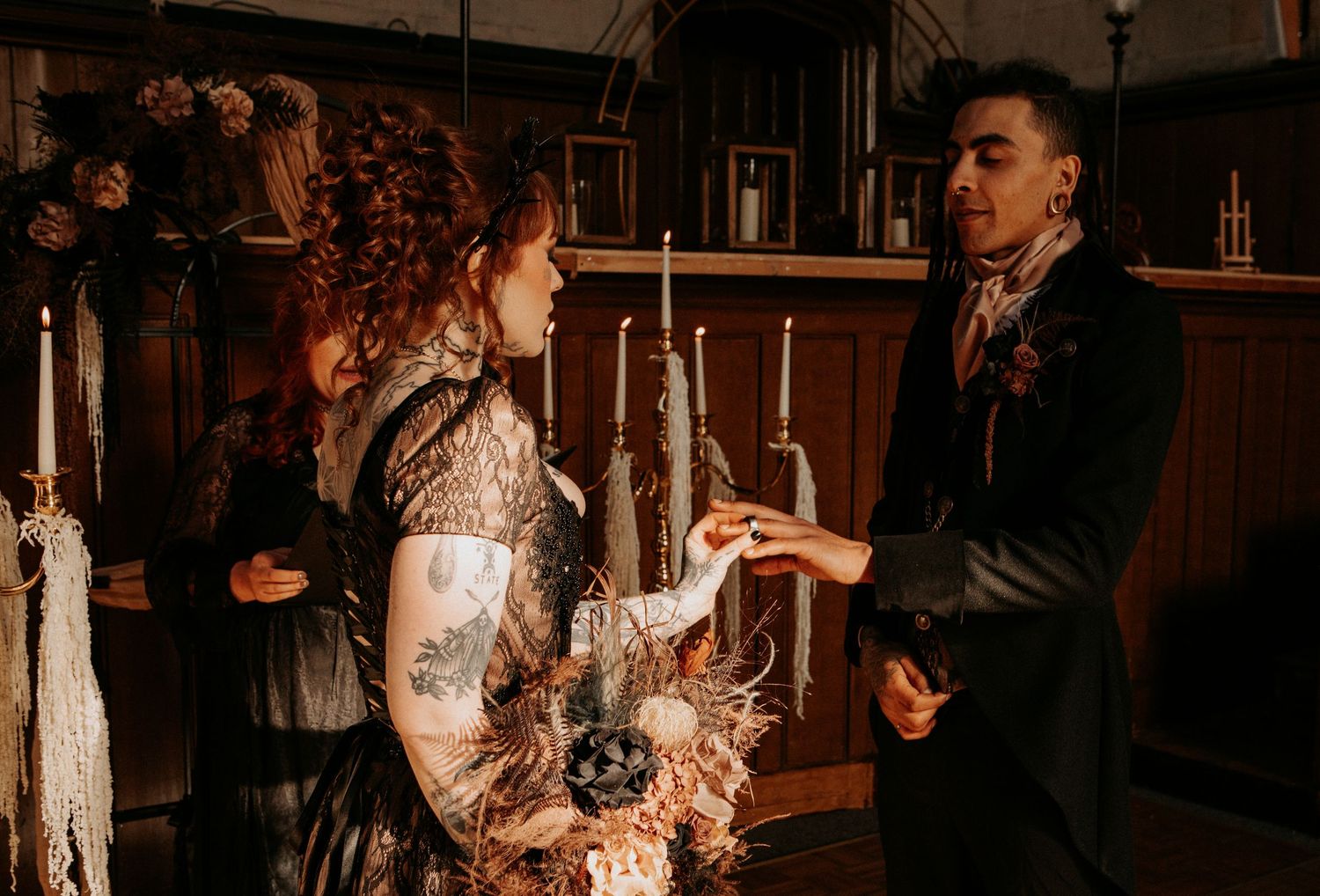 Gothic wedding ceremony with bride in black lace gown and groom exchanging rings in a candlelit venue.