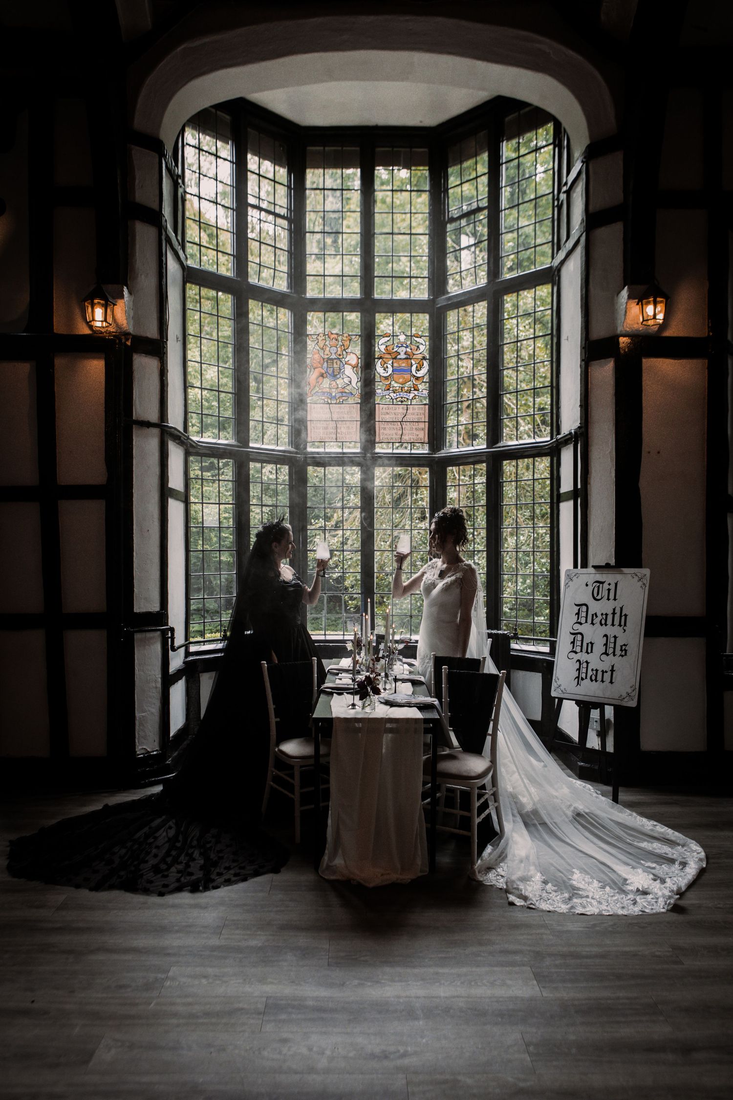 Two brides in black and white gowns stand at a sweetheart table before a grand stained glass window in a gothic wedding.