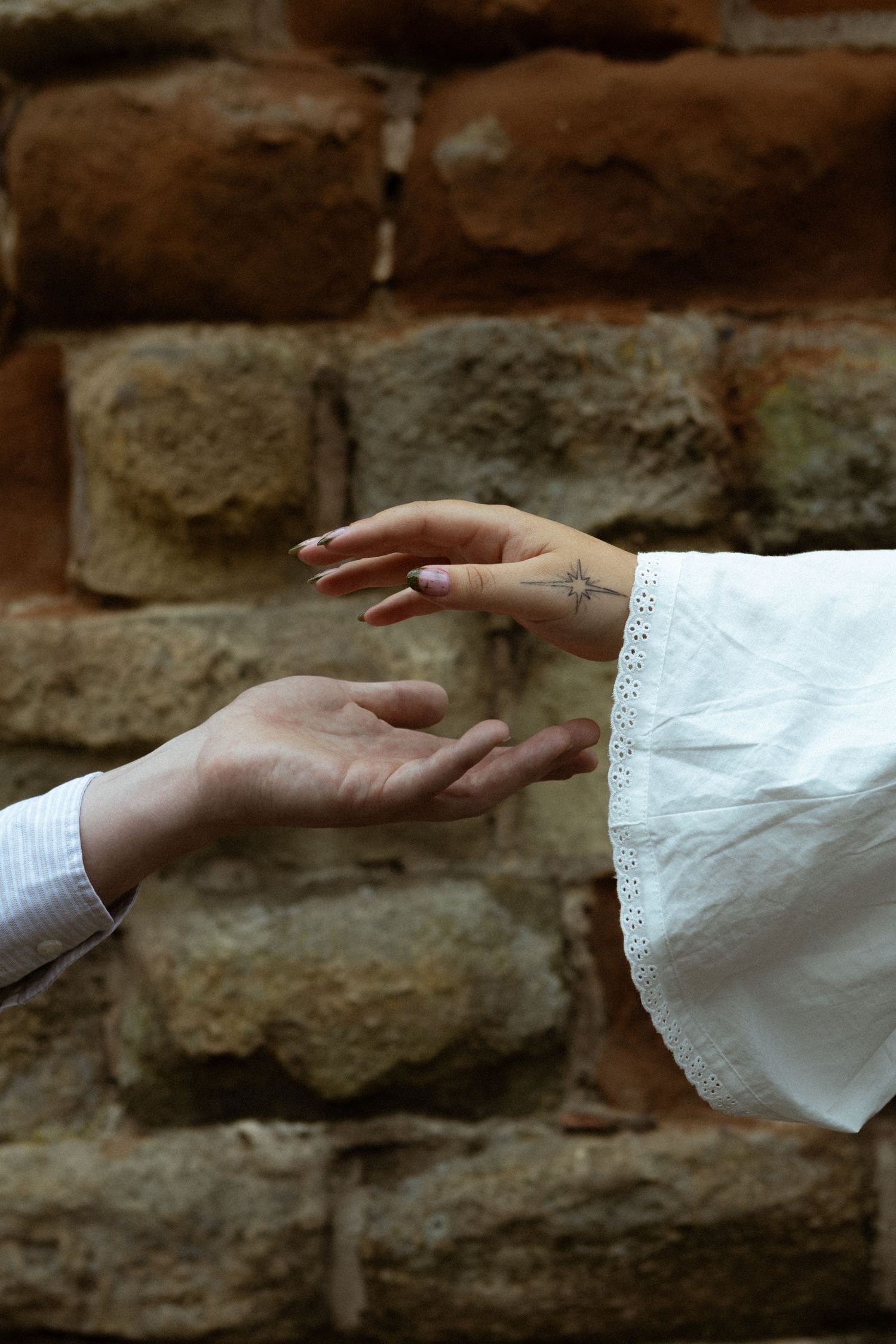 Two hands reaching toward each other against a rustic brick wall, one wearing a white lace sleeve with a small cross tattoo visible.