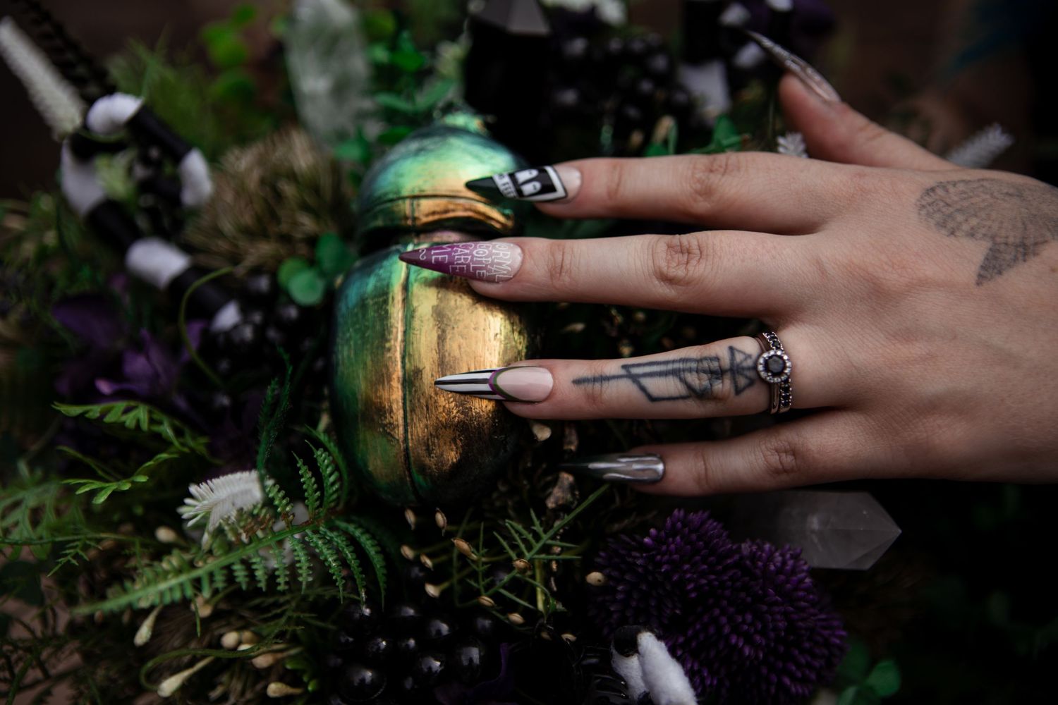 Hand with geometric nail art and tattoo resting on iridescent beetle amid dark floral arrangement with ferns.