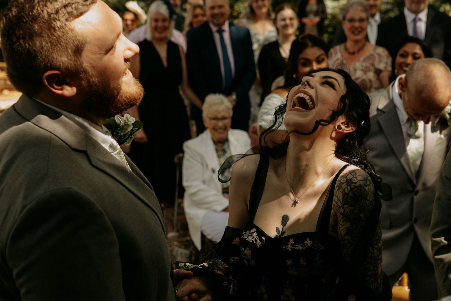 Bride in black lace dress laughs joyfully with groom at wedding ceremony surrounded by guests.