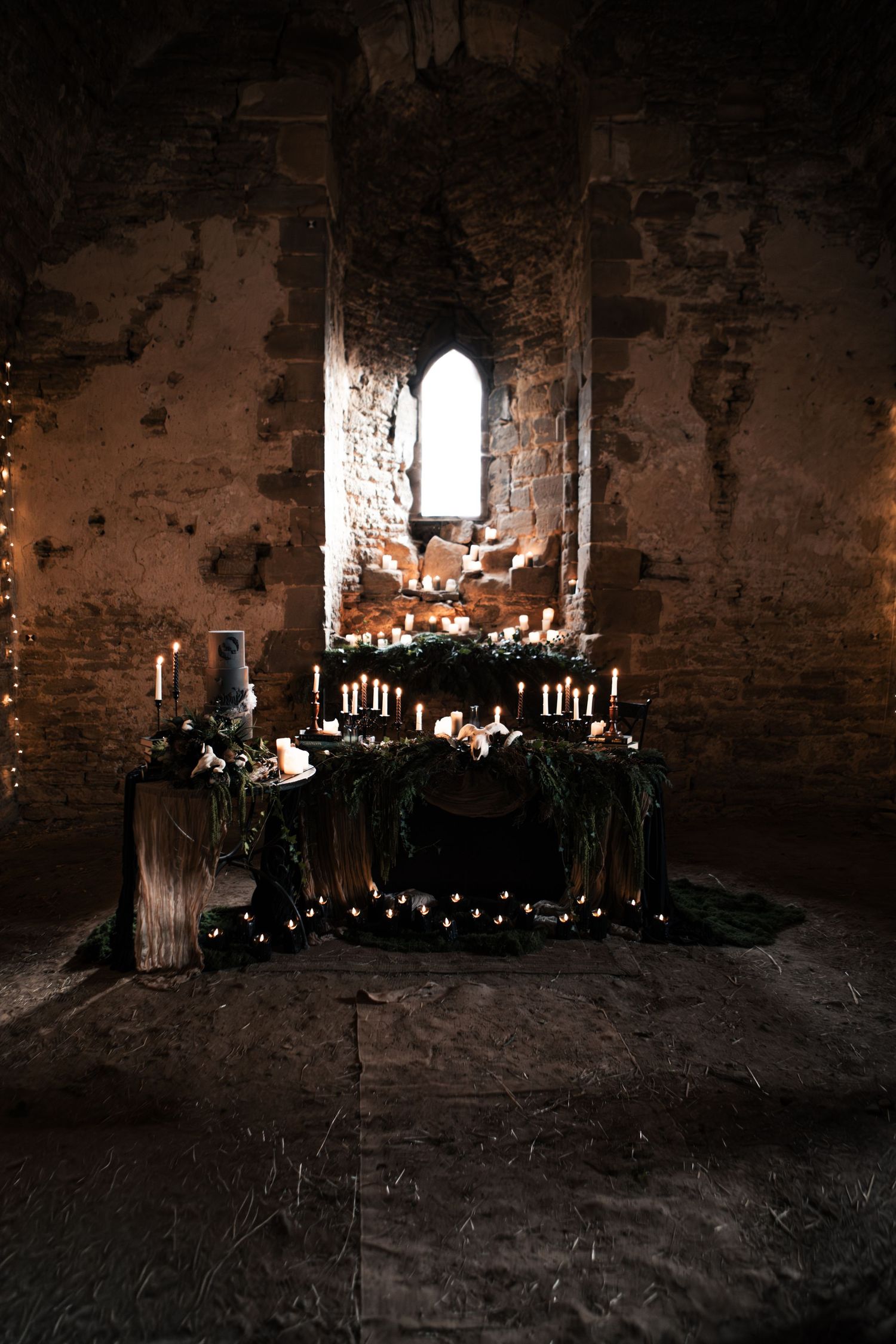 Gothic wedding altar in ancient stone ruins with candles, greenery, and arched window backdrop.
