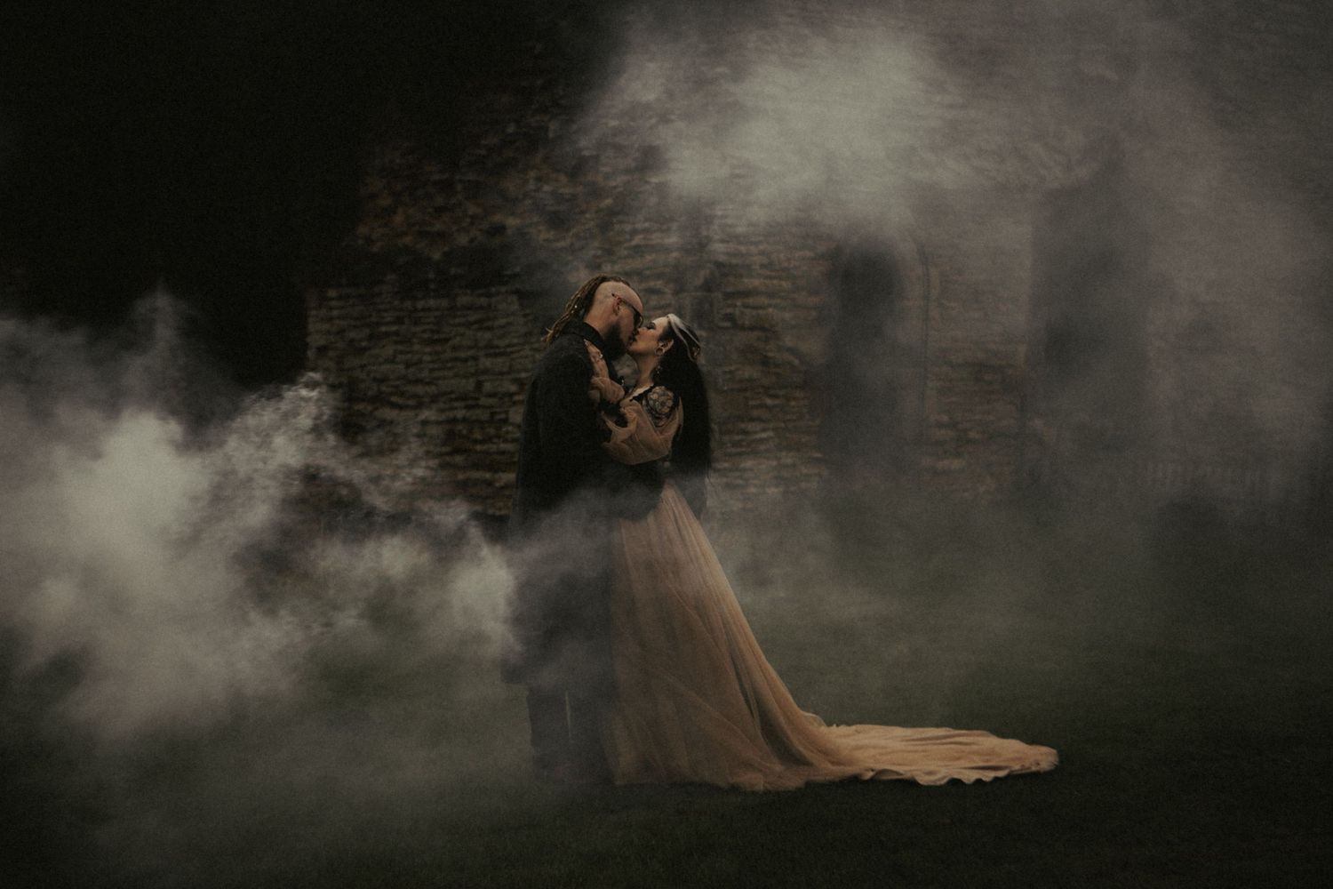 A couple embraces romantically in a dramatic fog-filled dark setting, with the bride in a flowing gown near a stone wall.
