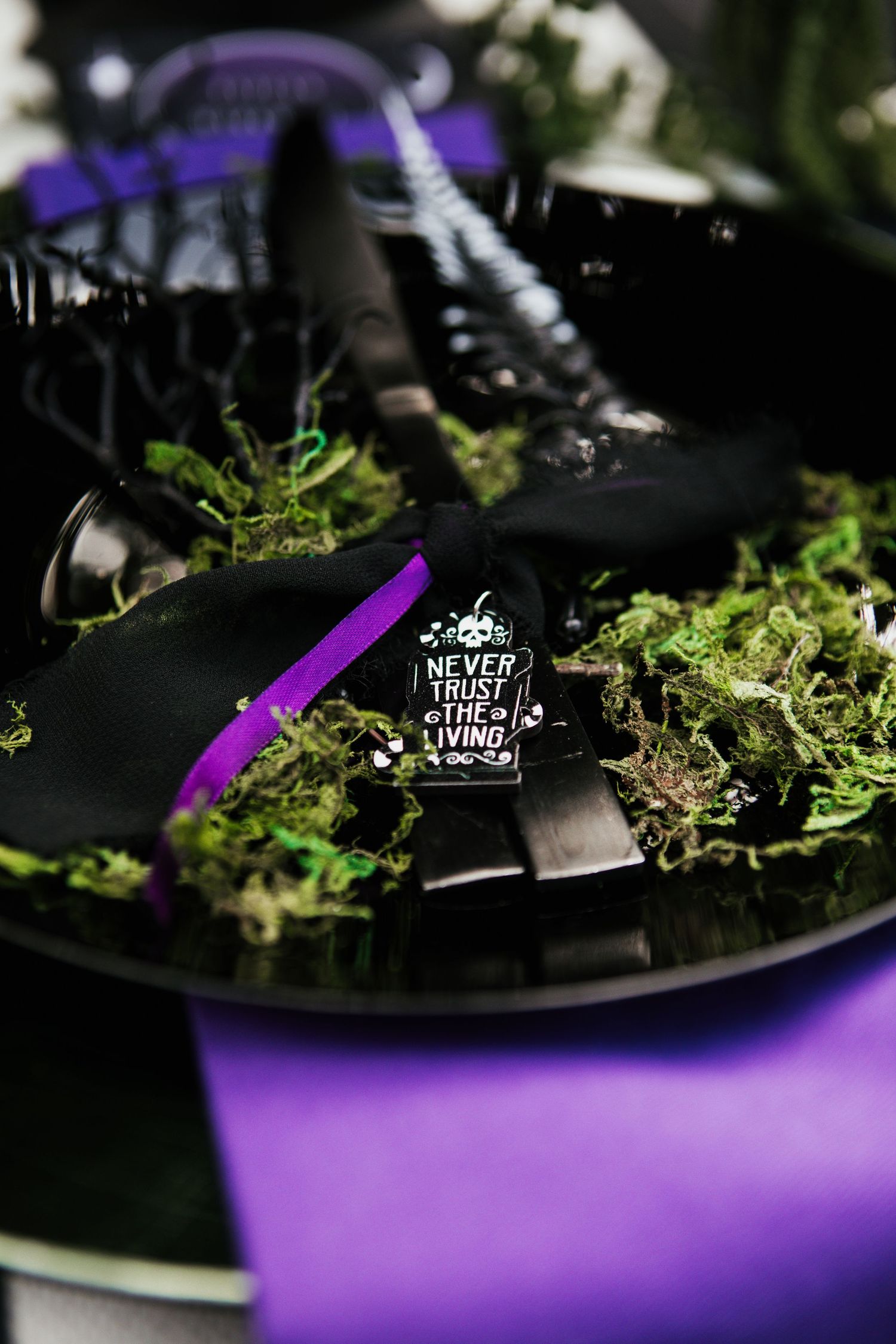 Black platform boots with a purple ribbon and a 'Never Trust the Living' enamel pin displayed on green moss.