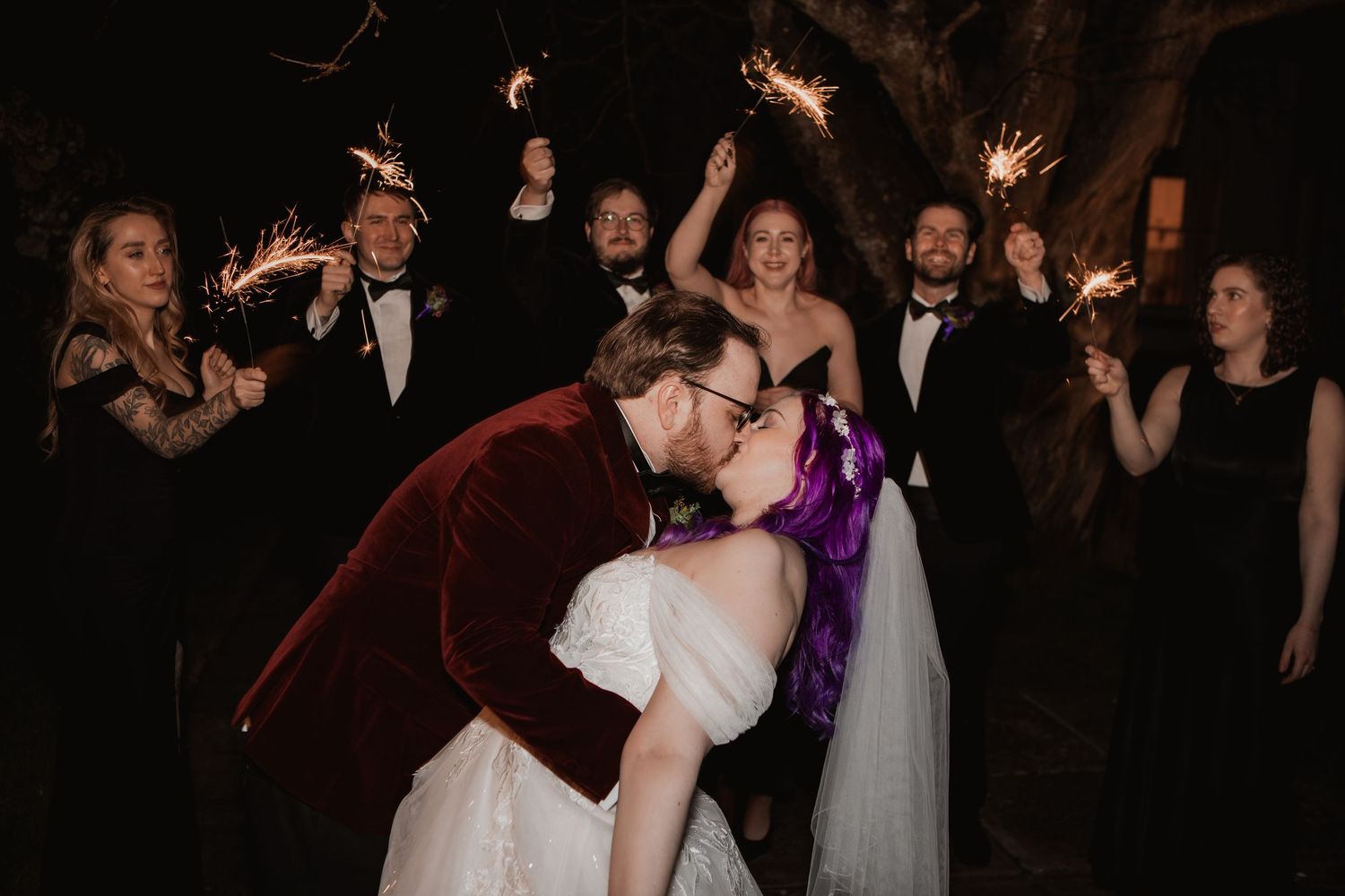 Bride and groom share a kiss surrounded by wedding party holding sparklers at night outdoor celebration.
