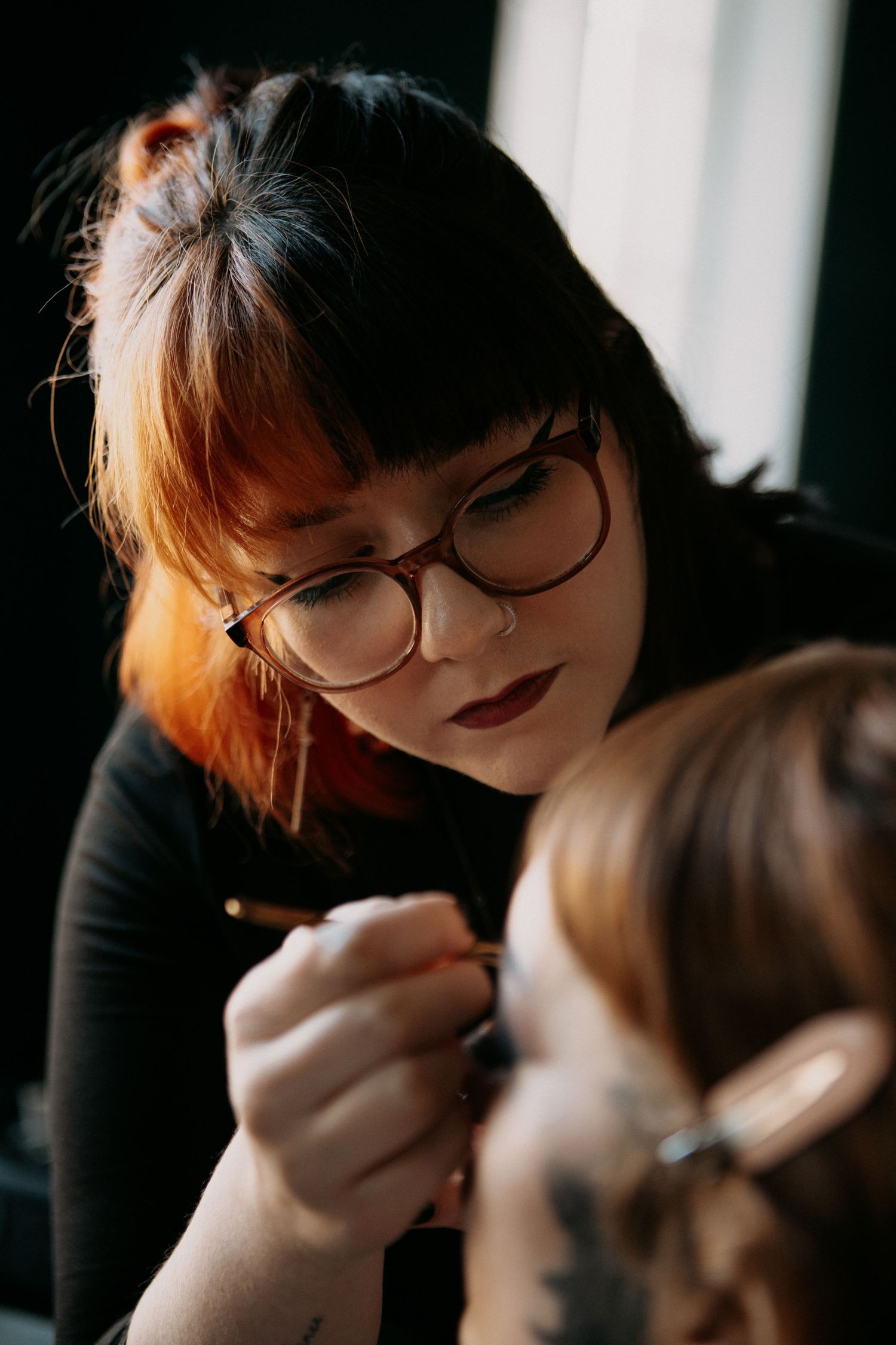 Professional makeup artist with red-tinted hair and glasses carefully applying makeup to a client.