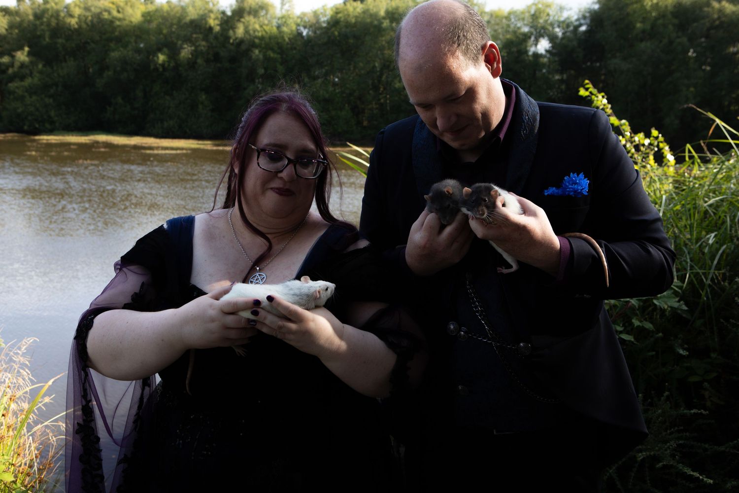 Couple in dark wedding attire holding ducklings beside a river in outdoor nature setting.