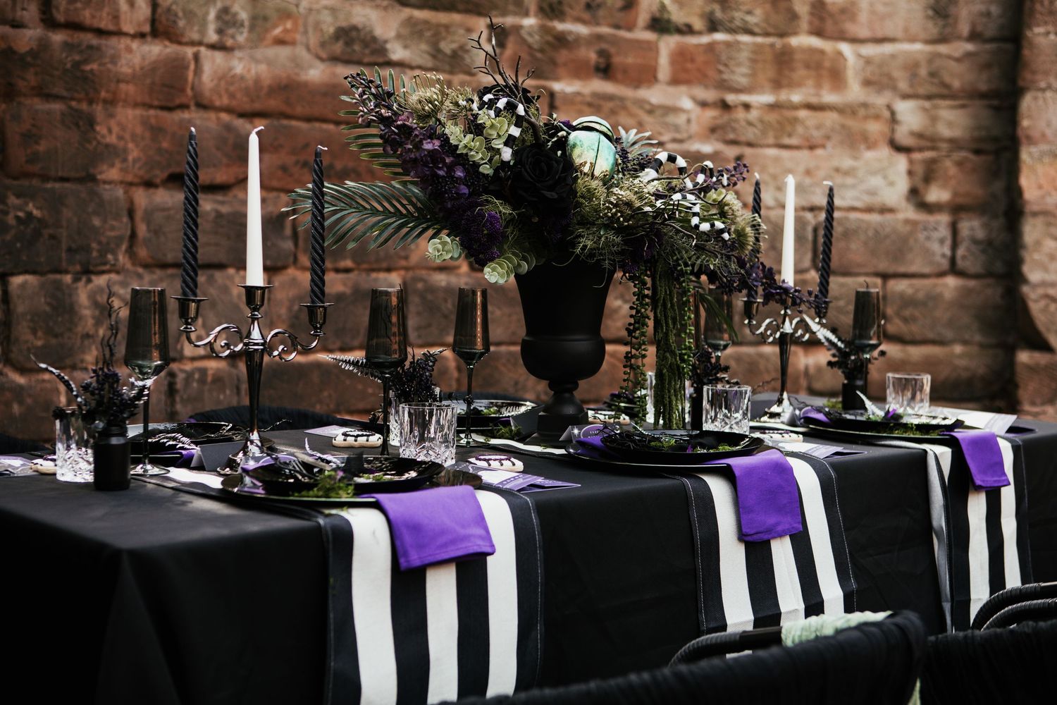 Gothic wedding tablescape with black linens, purple napkins, black candlesticks, and dark floral centerpiece against brick wall.