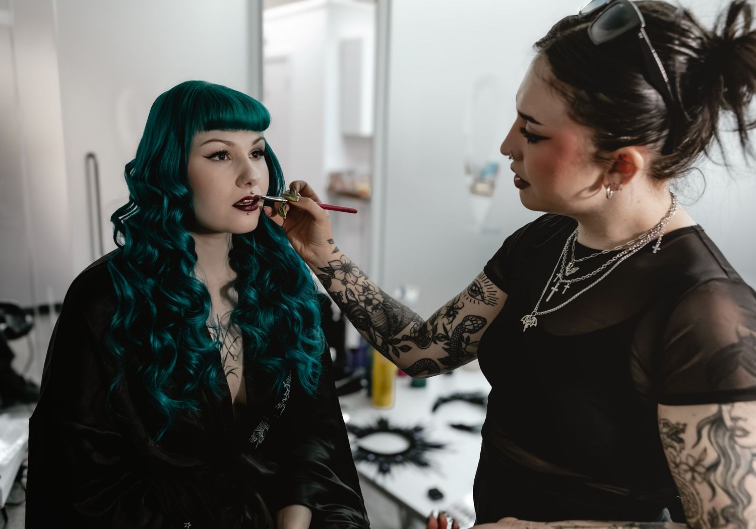 Tattooed makeup artist applying lipstick to a woman with teal wavy hair and bangs in a bright studio.