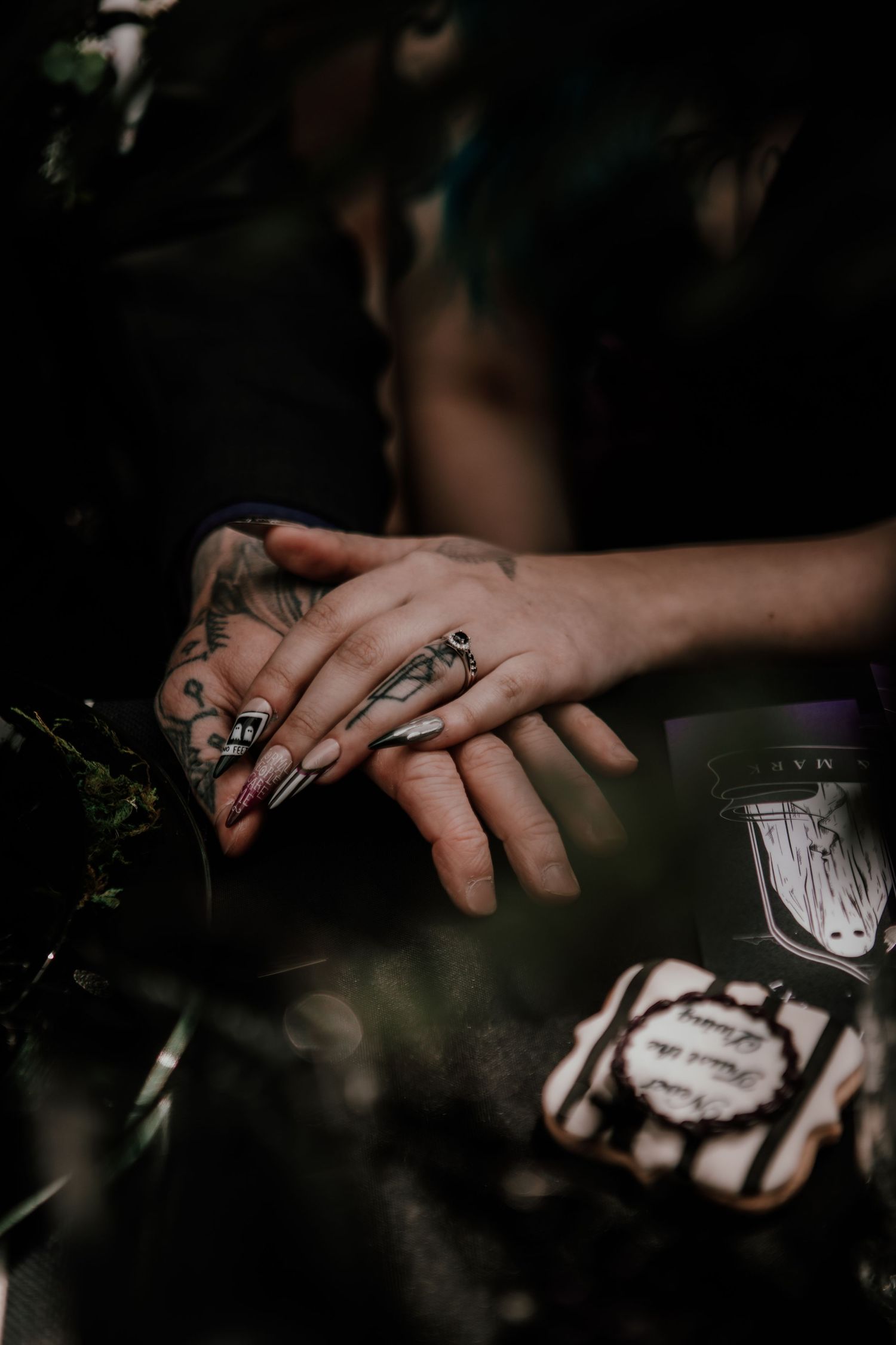 Tattooed hands with dark nail polish and rings resting together in a moody, gothic-inspired close-up shot.