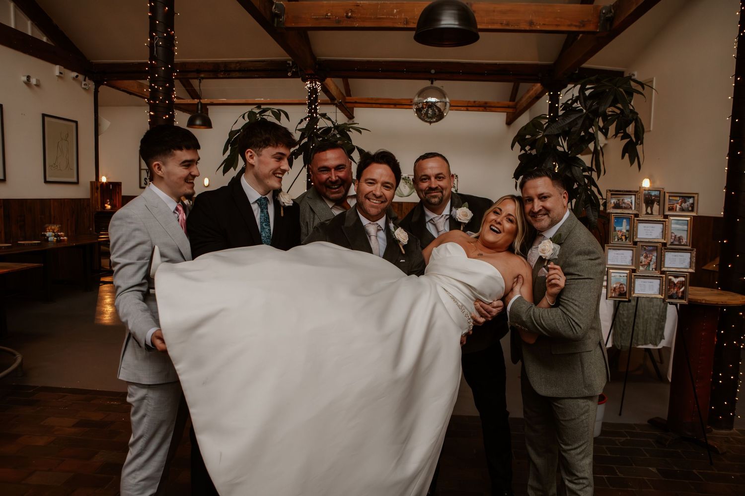 A bride in a white dress is being lifted and carried by a group of groomsmen in suits at an indoor reception venue.