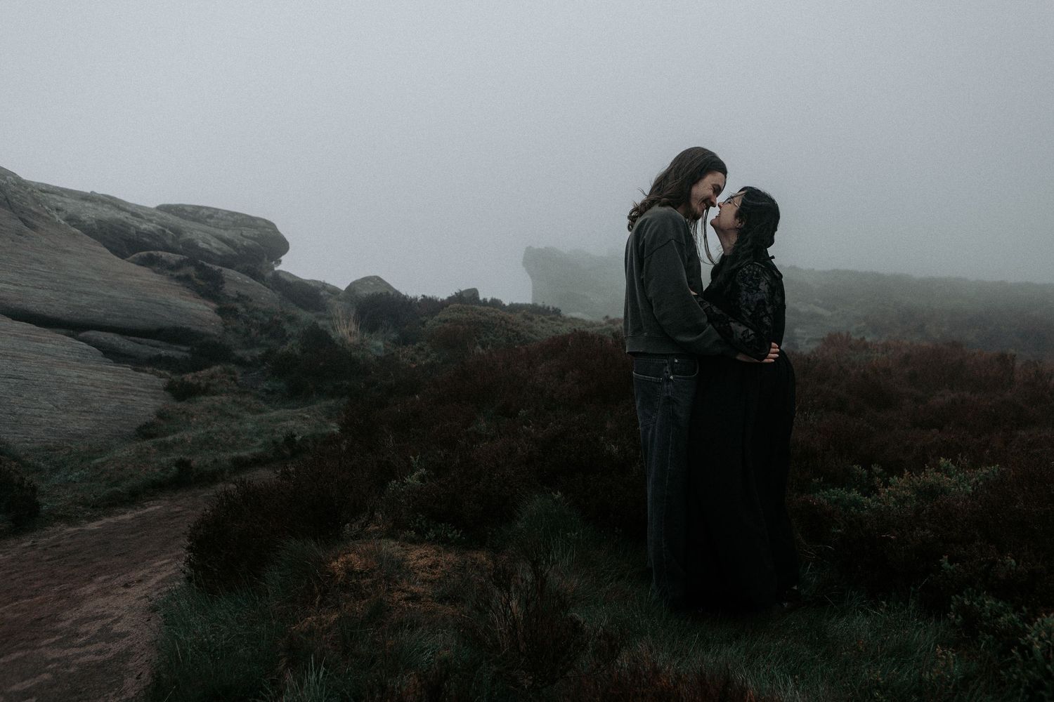 A couple stands facing each other tenderly in a foggy moorland landscape with rocky outcrops.