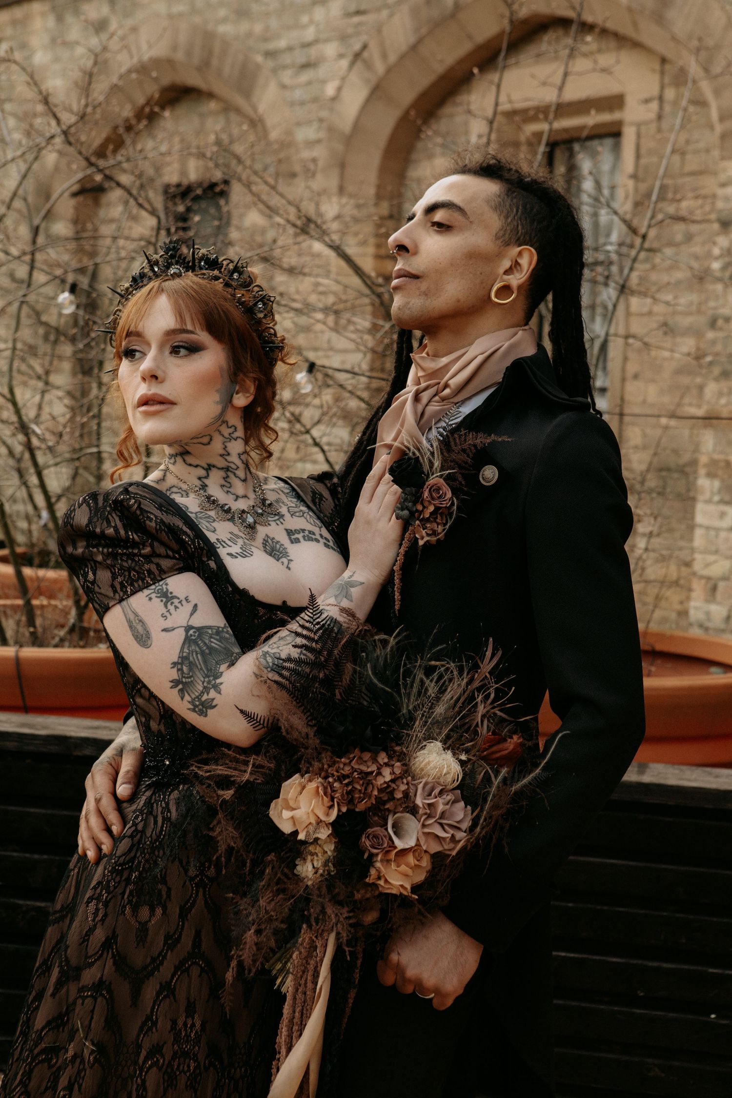 Gothic wedding couple in black attire with tattoos and dark floral bouquet posing at a stone building.