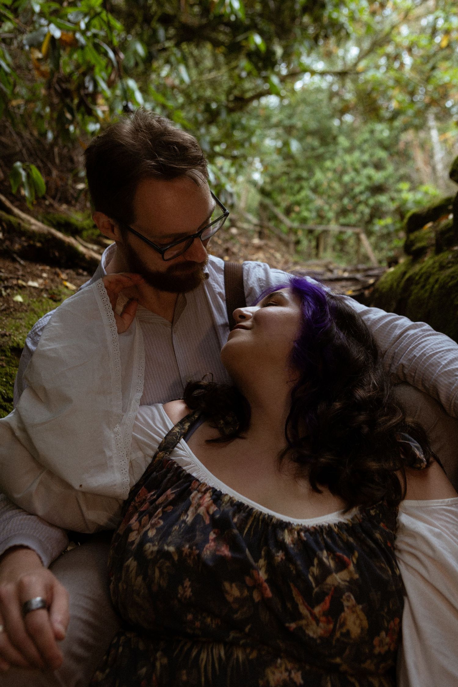A couple shares a romantic moment in a lush forest, woman with purple hair leaning back as man in glasses gazes at her.
