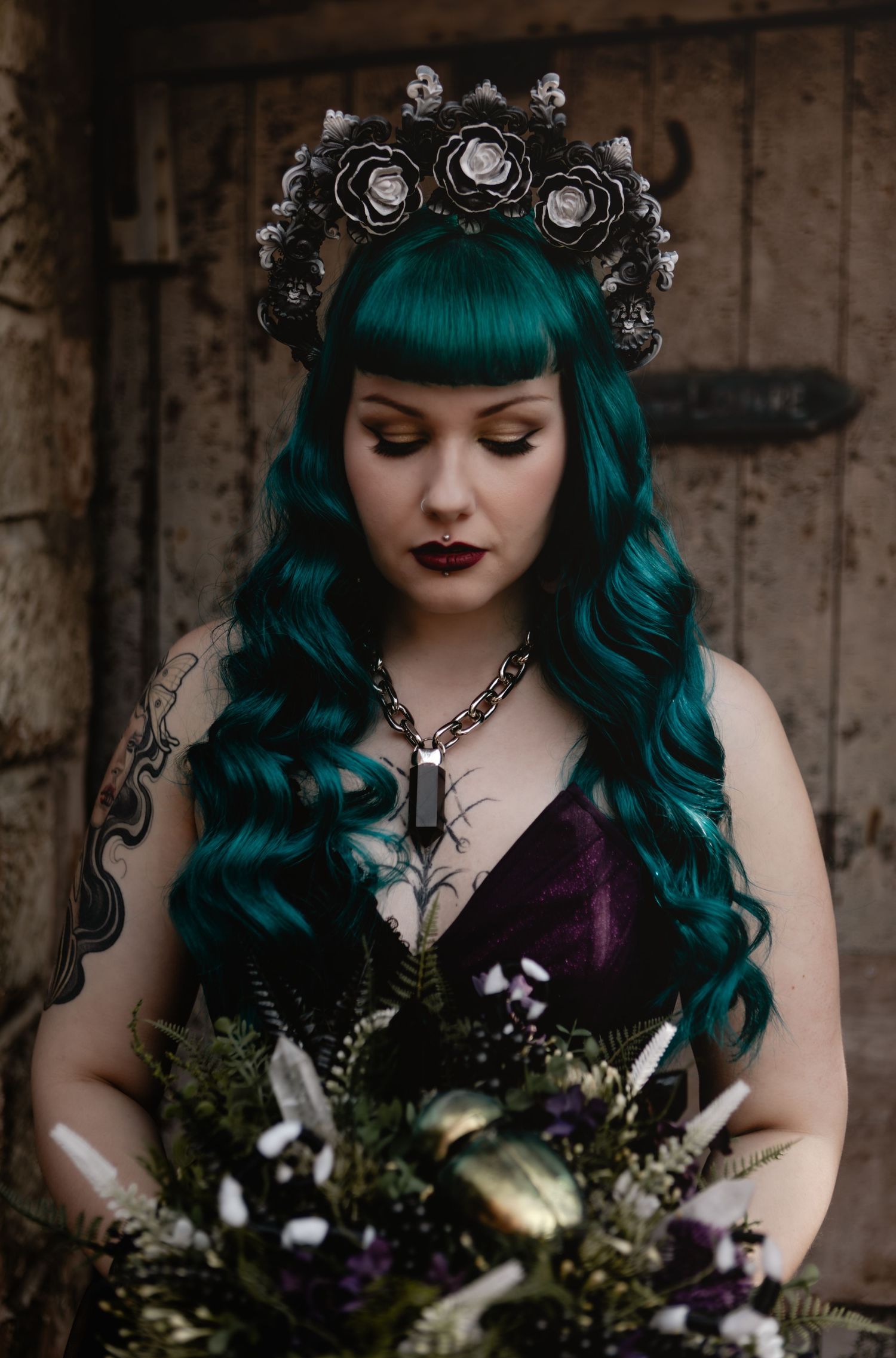 Gothic bride with teal wavy hair wears black floral crown and purple corset holding dark wedding bouquet.