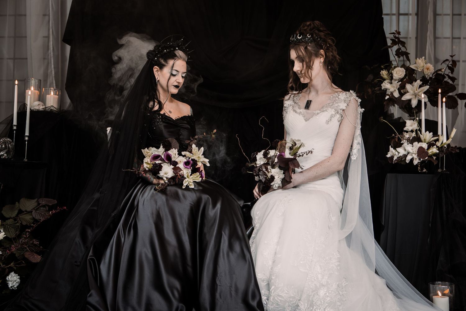 Two brides in black and white wedding gowns holding bouquets, seated together with candles and floral backdrop.