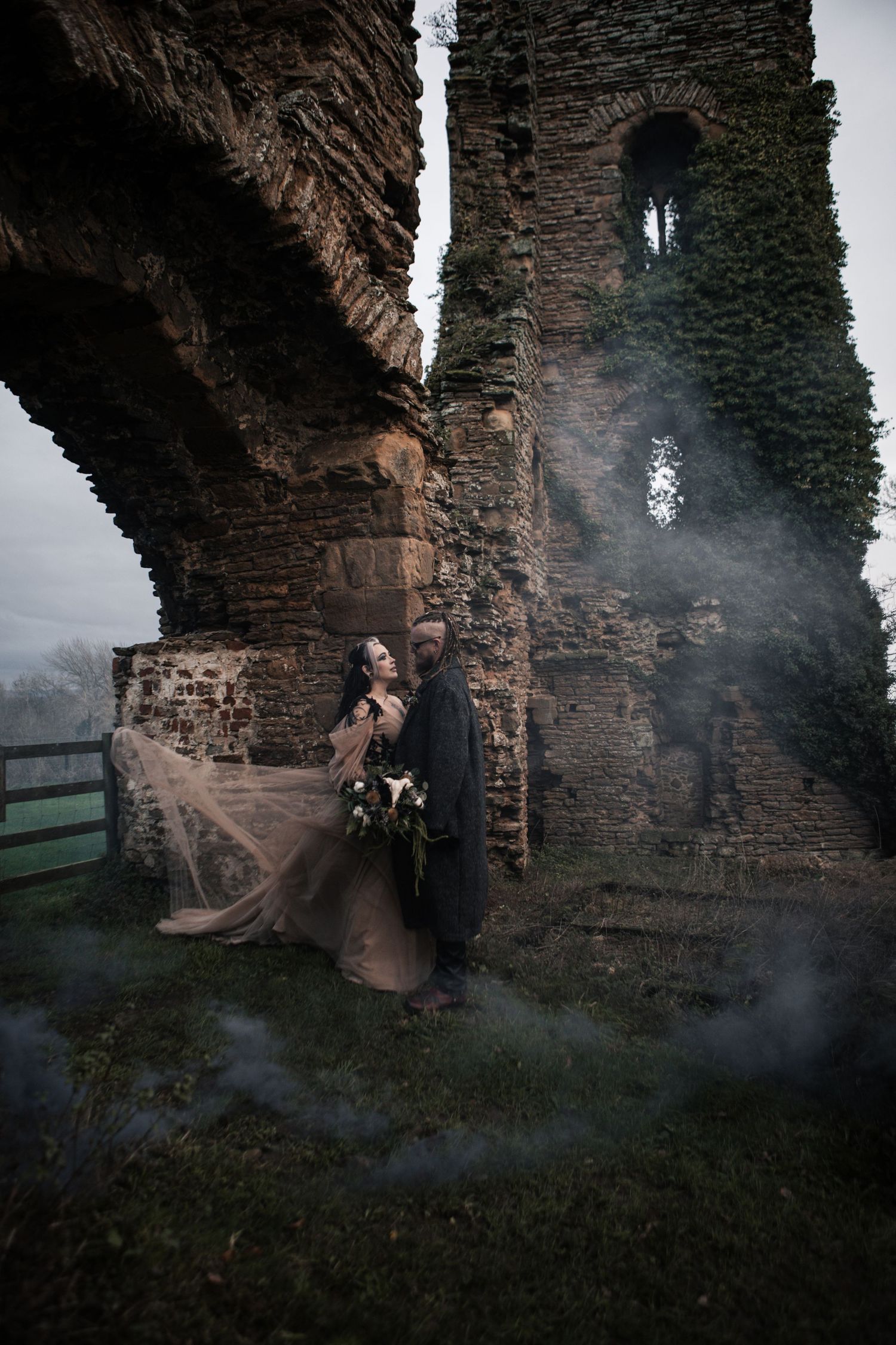A couple poses dramatically among ancient stone ruins with moody smoke effects for a gothic wedding.