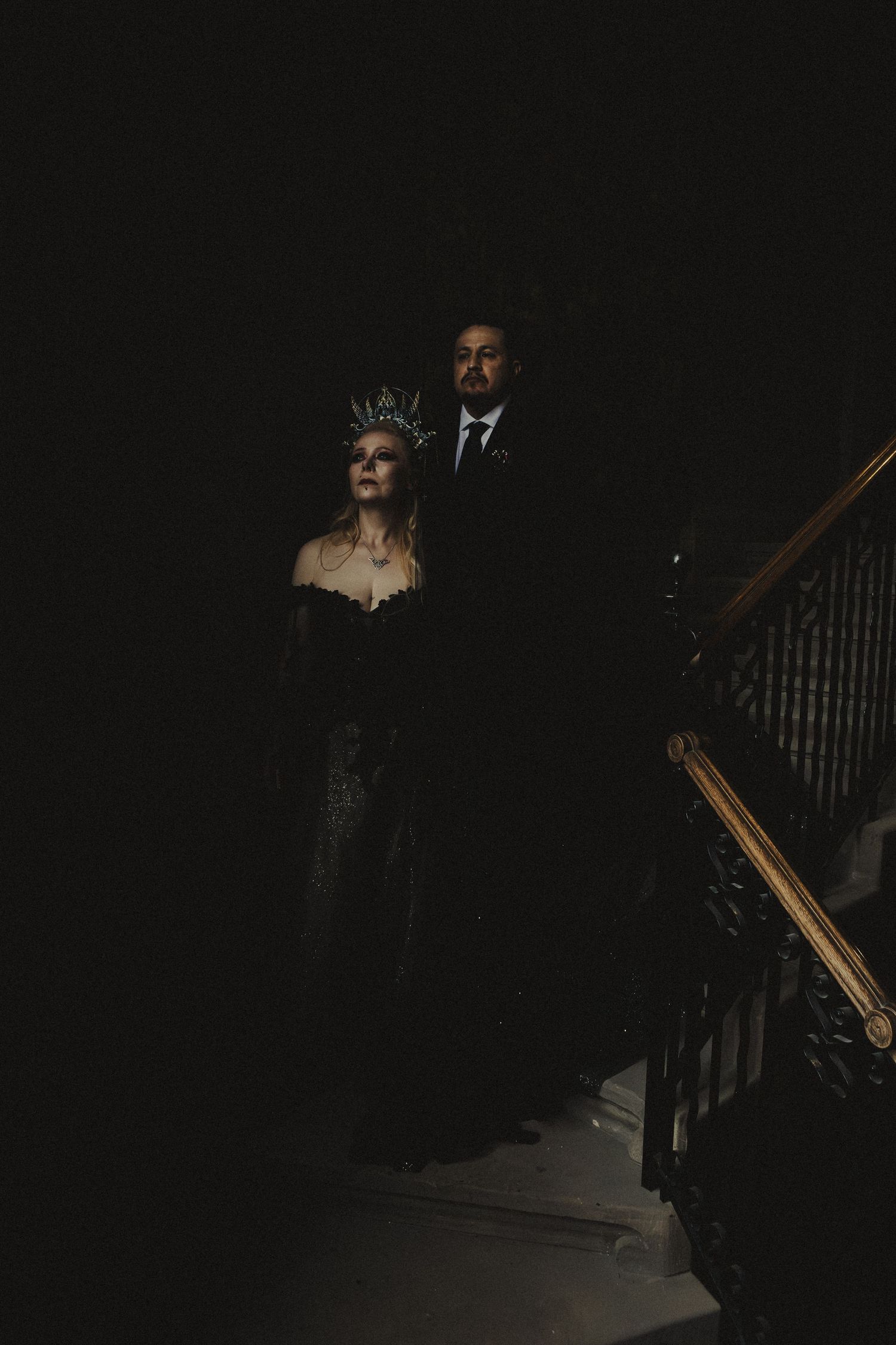 Gothic bride in black gown and crown with groom on dark staircase for dramatic Halloween wedding portrait.