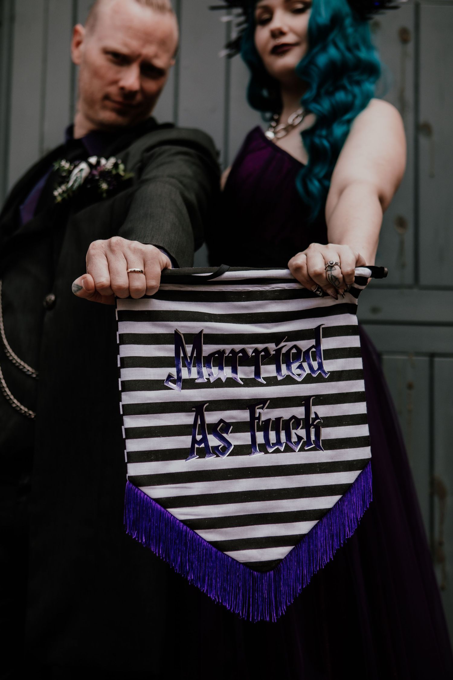 Couple holding a black and white striped pennant banner reading Married As Fuck with purple fringe at a gothic wedding.