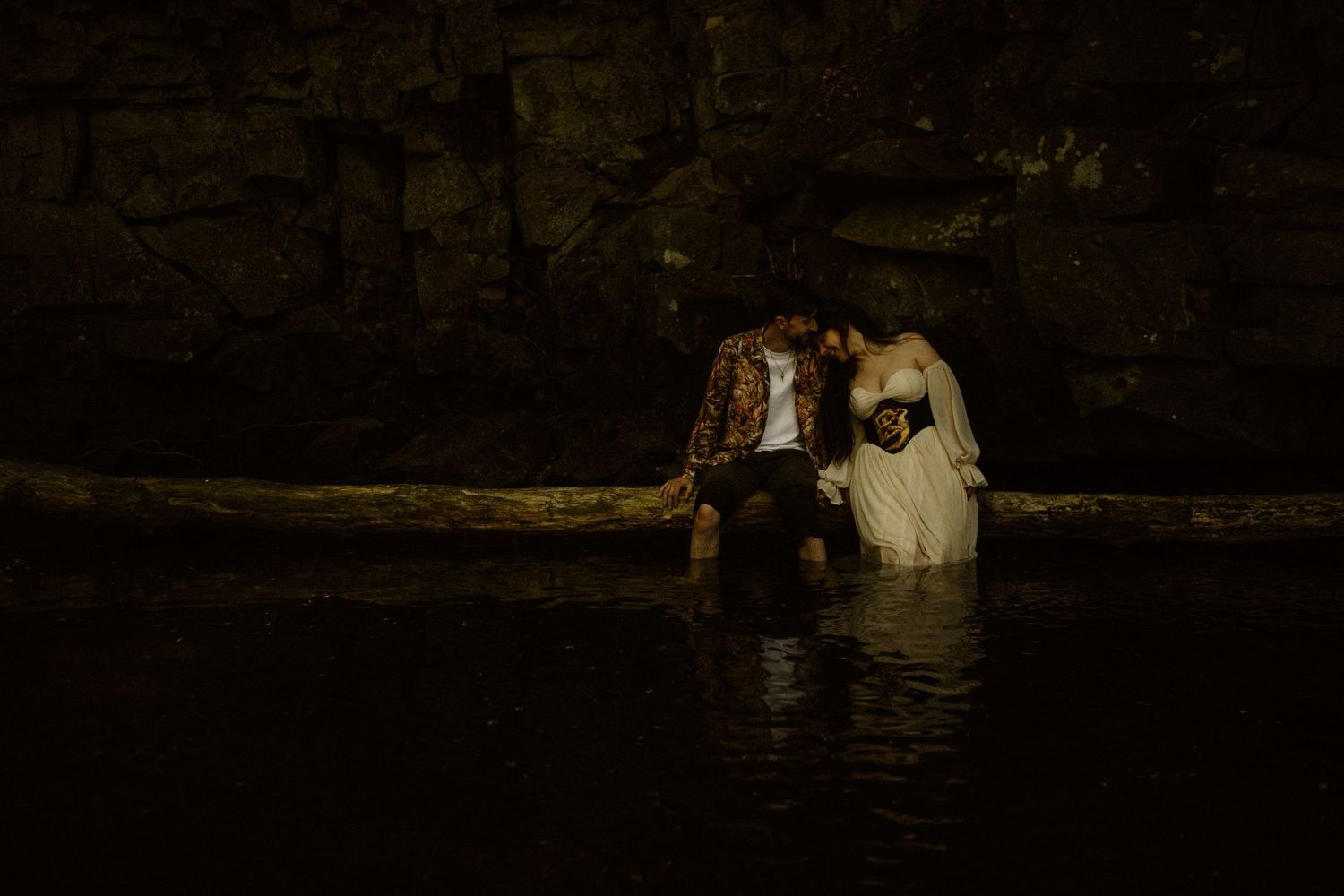 Couple in wedding attire sitting together in dark atmospheric cave or grotto setting.