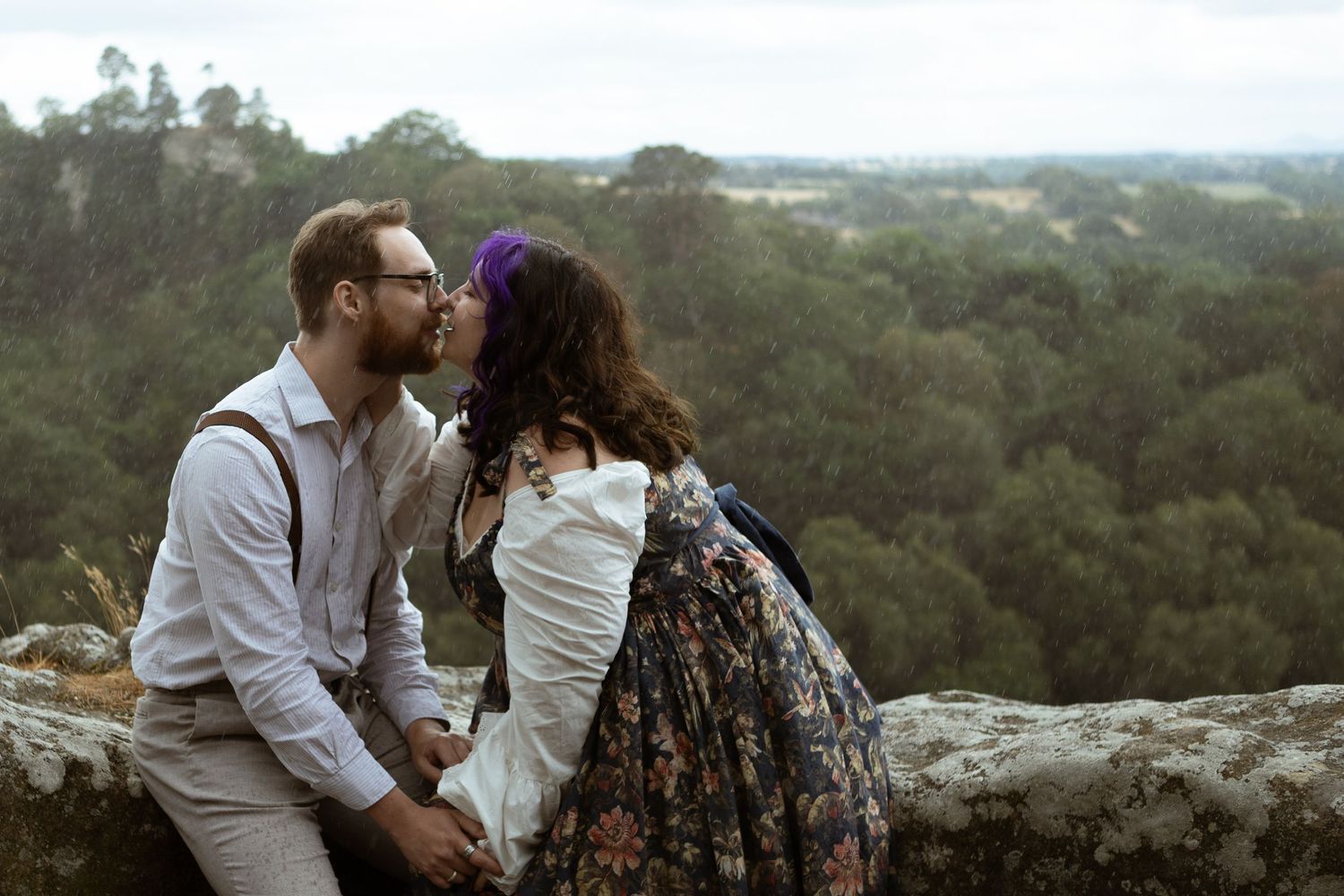 A couple shares a romantic kiss on a stone wall overlook surrounded by lush green forest landscape.