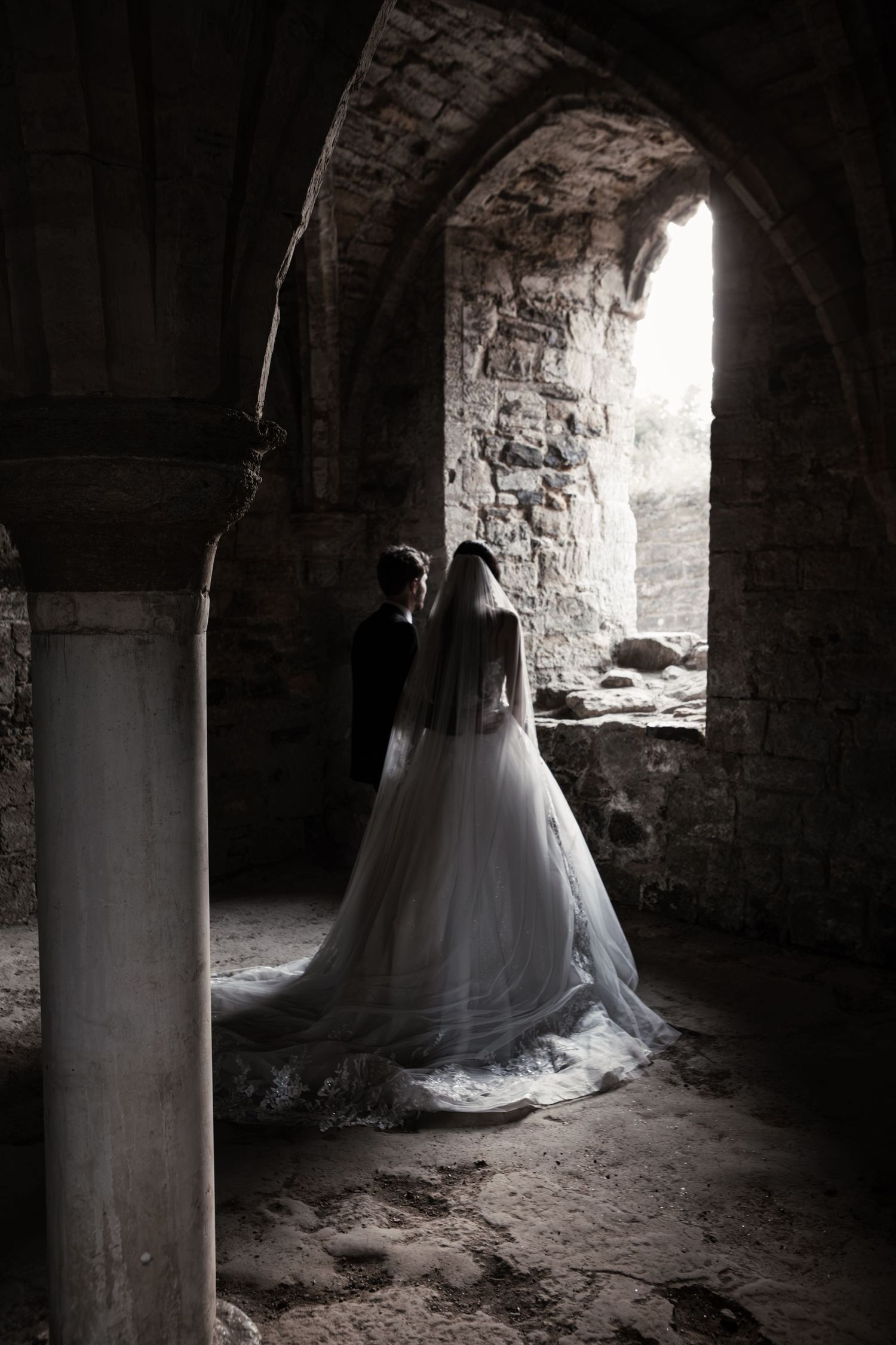 Bride and groom standing in a dramatic stone archway of ancient ruins, bride wearing a flowing white gown with long veil.