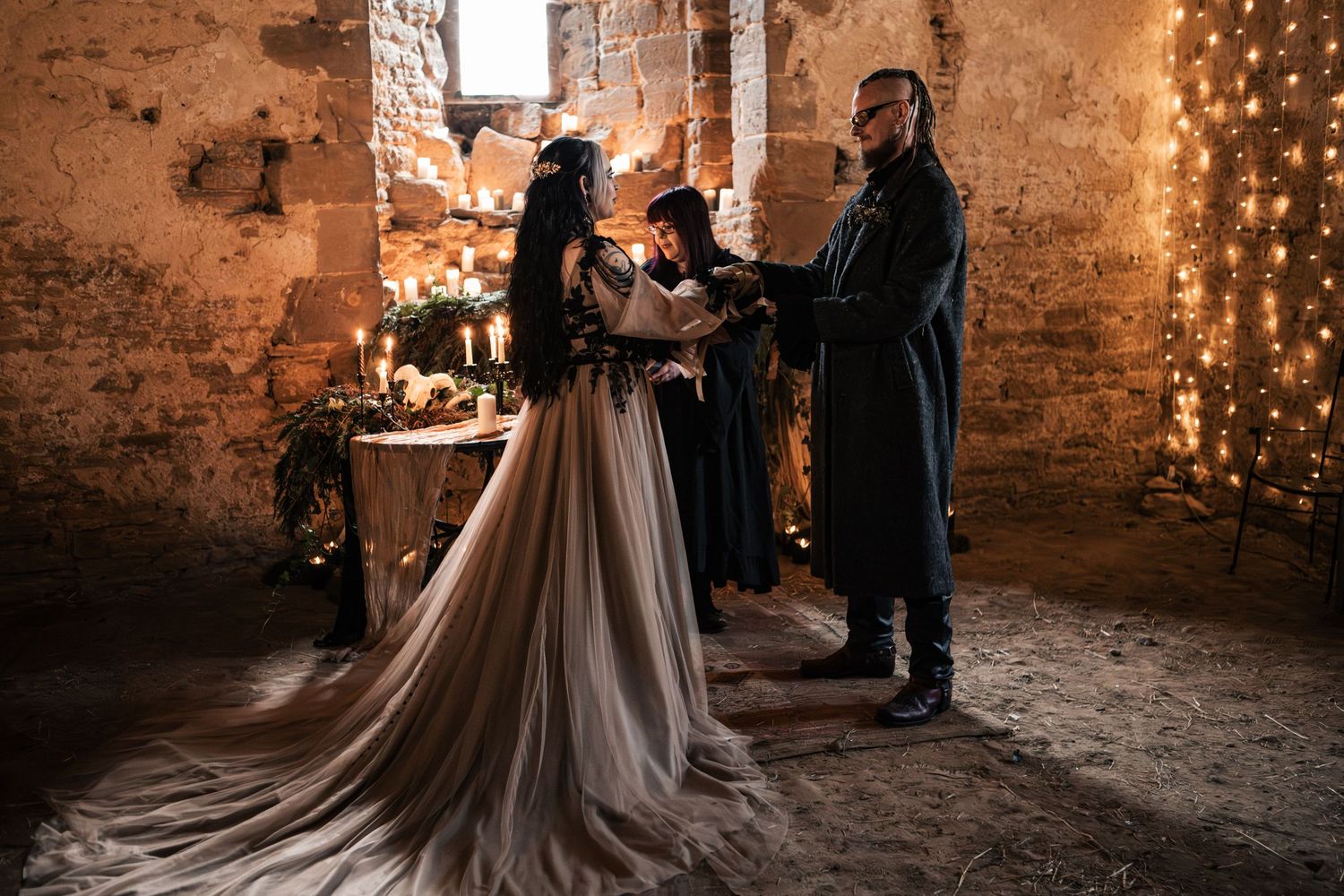 Gothic wedding ceremony in candlelit stone ruins, bride in flowing gown and groom in dark coat exchanging vows.