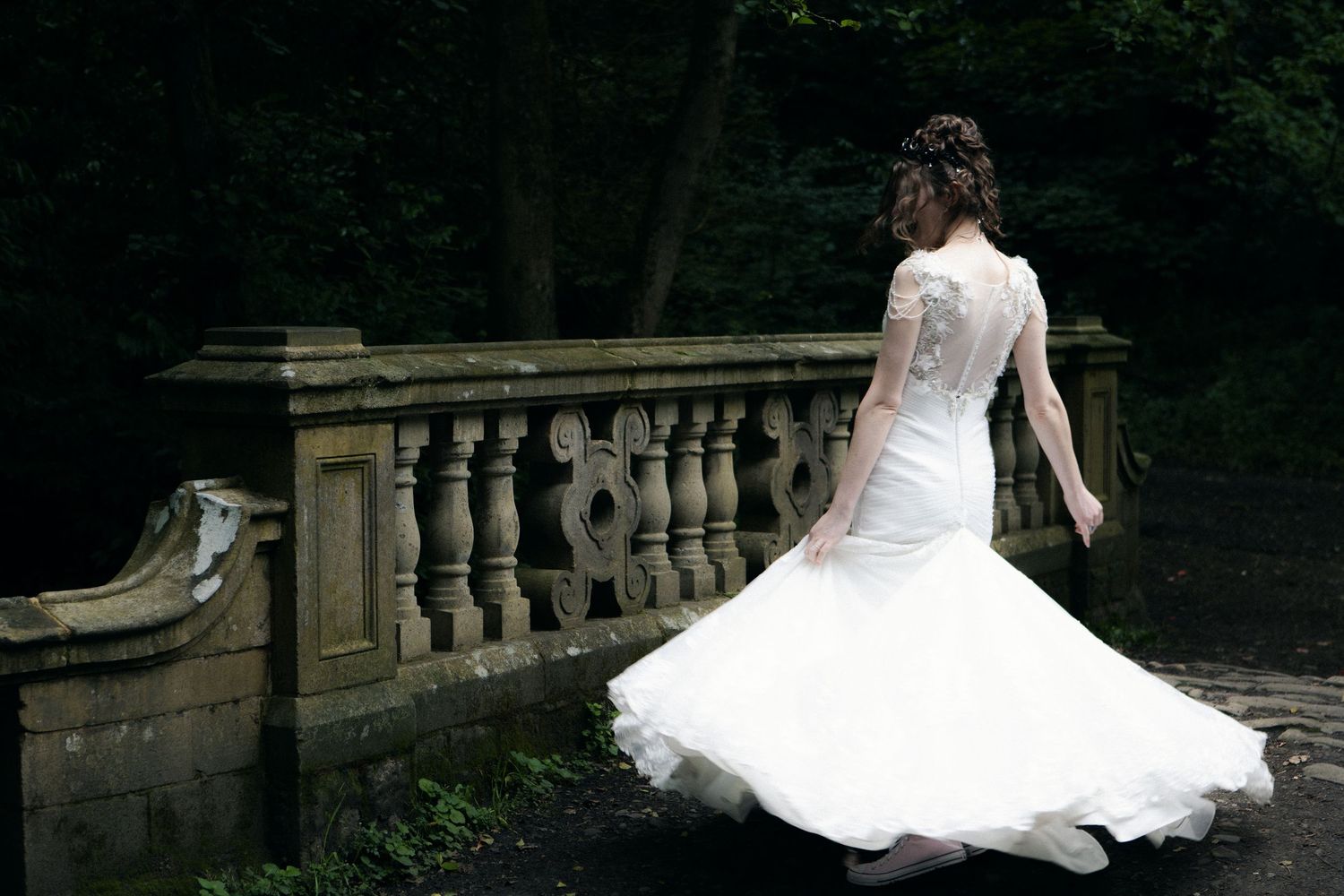 Bride in white mermaid wedding dress twirling on stone balustrade bridge surrounded by lush green trees.