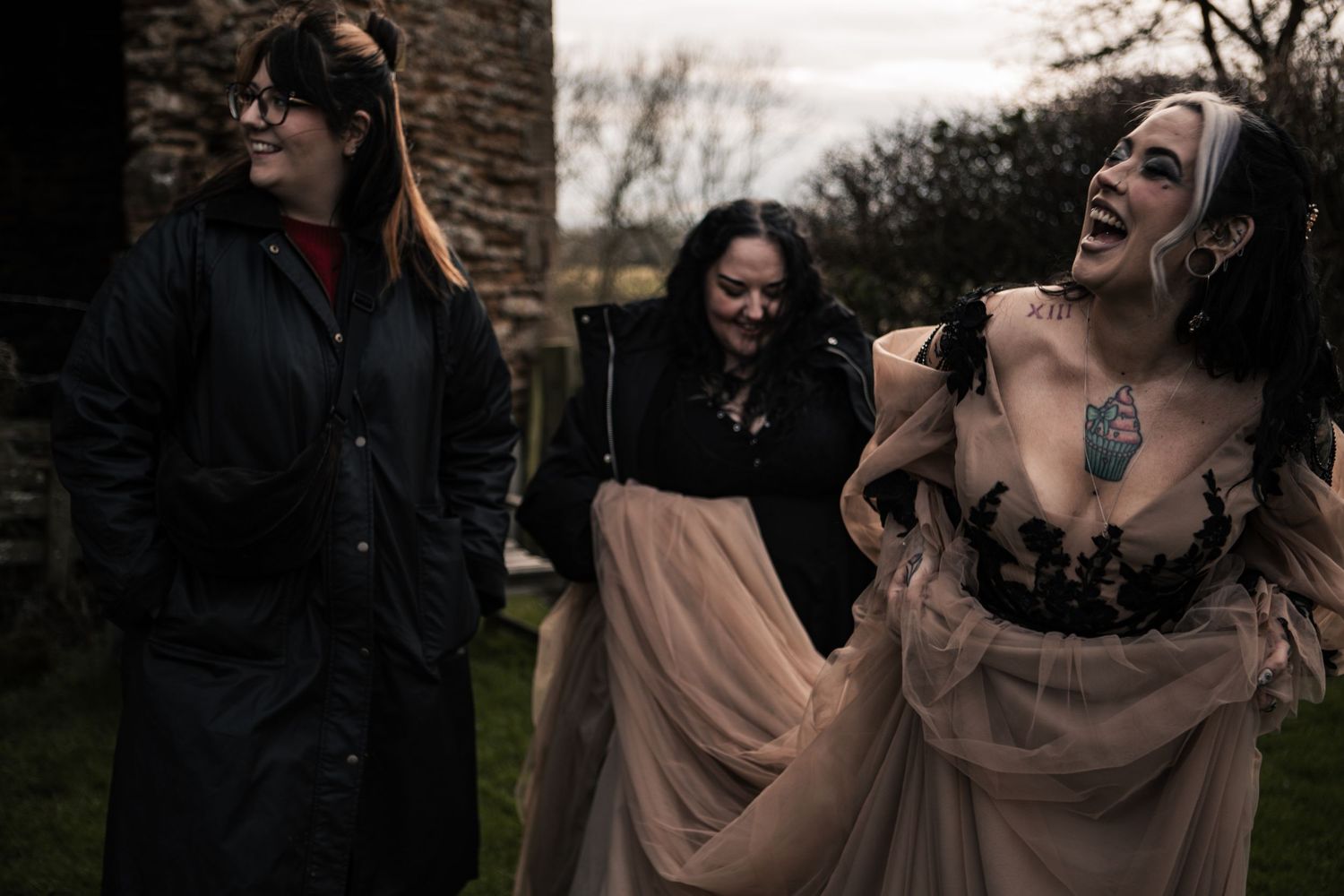 Three women laughing outdoors near stone ruins, one wearing a flowing blush tulle gown with black lace.