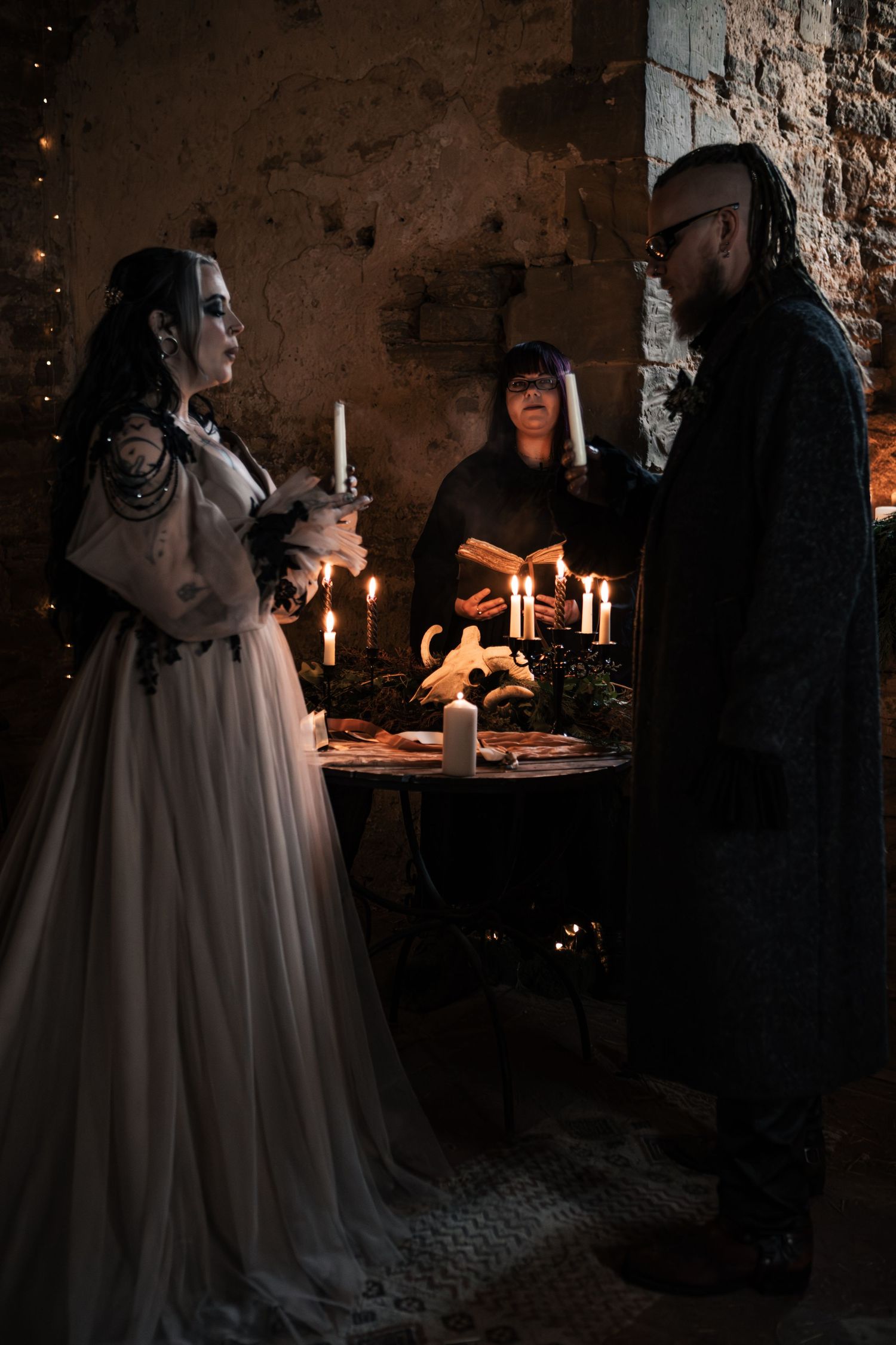 Gothic wedding ceremony with bride in white gown and groom in black coat exchanging vows by candlelight in stone ruins.